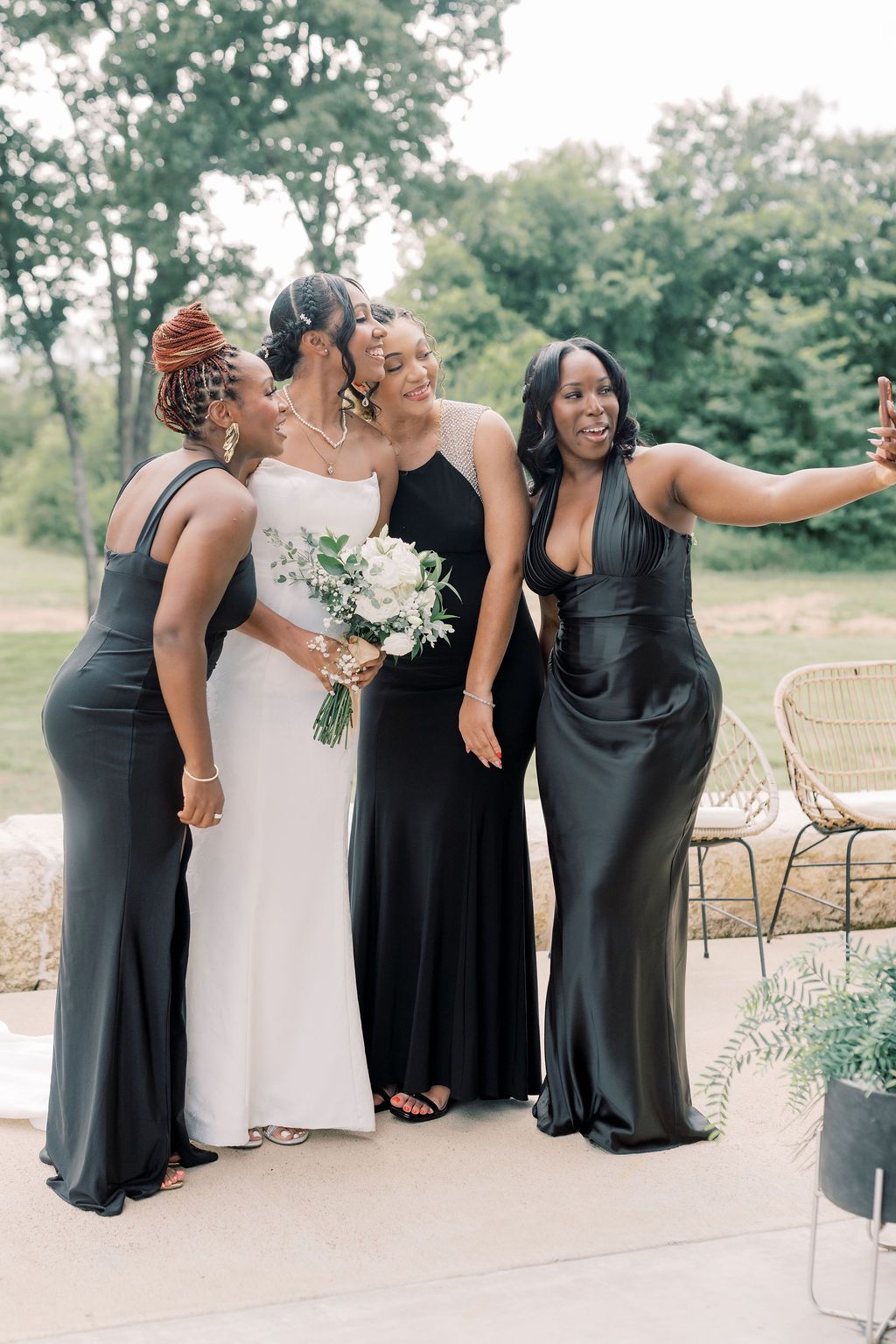 Bride in white gown poses with three bridesmaids in black dresses taking a selfie outdoors at a wedding.