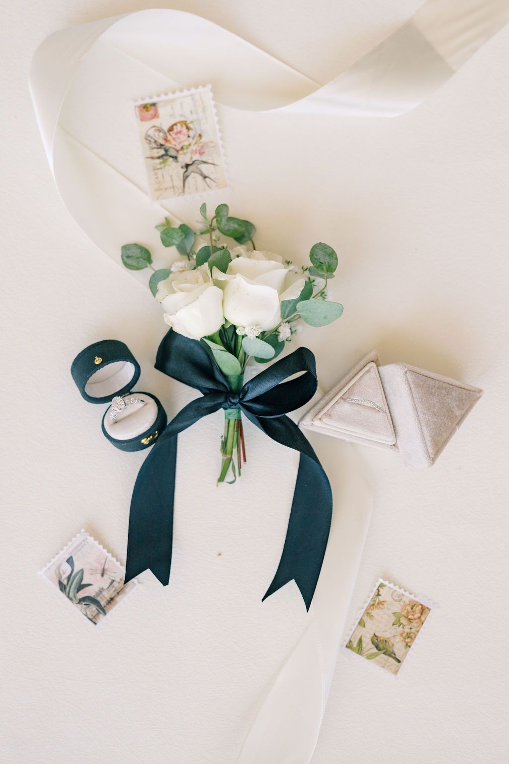 White rose boutonniere with dark green ribbon beside wedding rings, velvet box, and vintage stamps on cream background.