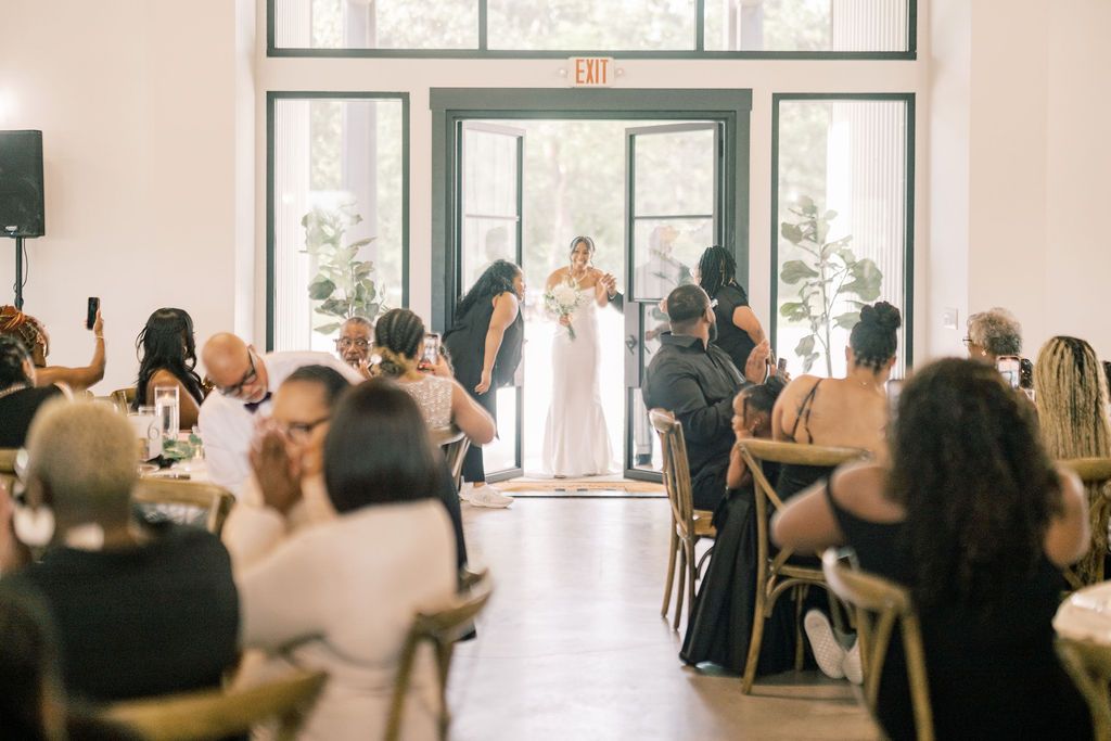 Elegant wedding ceremony in bright venue with bride at altar, guests seated in wooden chairs, large windows in background.