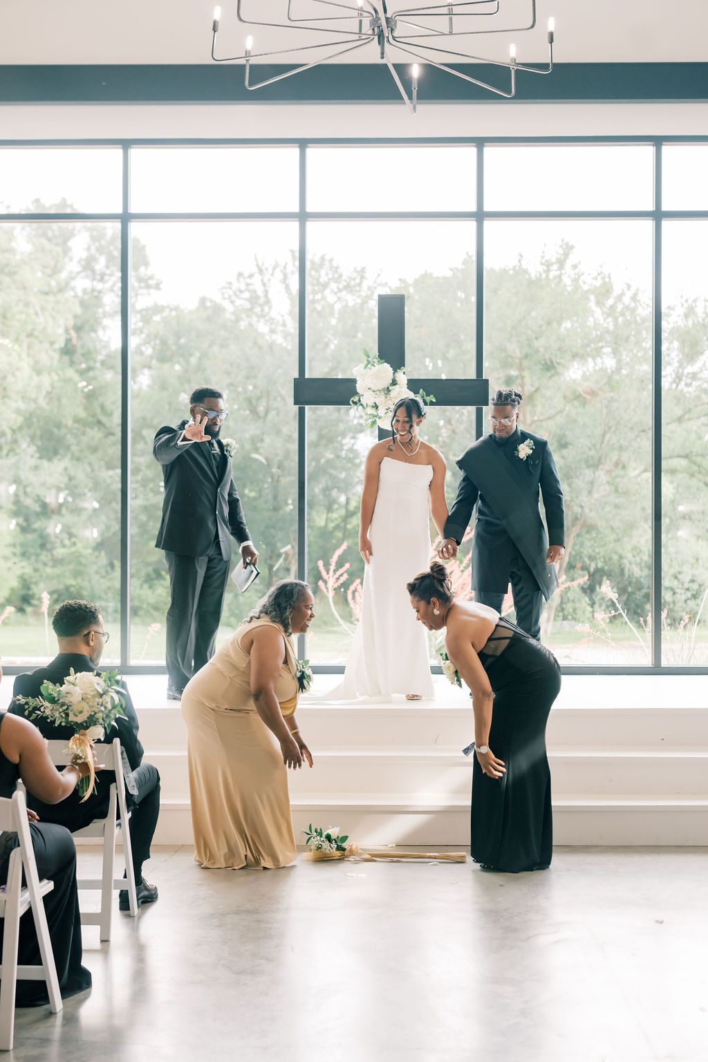 Bride stands at altar with cross backdrop as bridesmaids pick up bouquet during elegant indoor wedding ceremony.