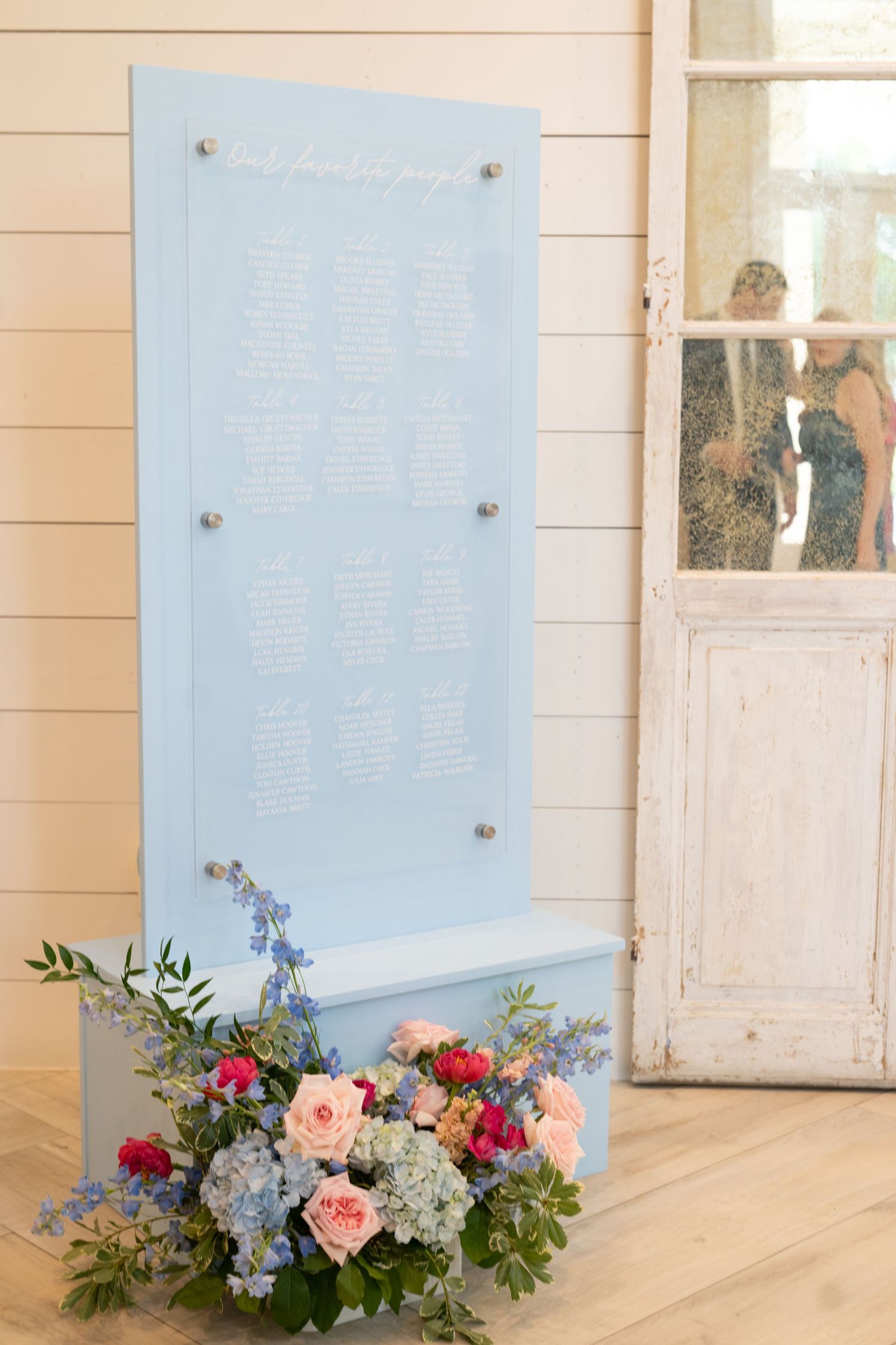 Light blue wedding seating chart display with pink and blue floral arrangement at base, elegant white shiplap backdrop.