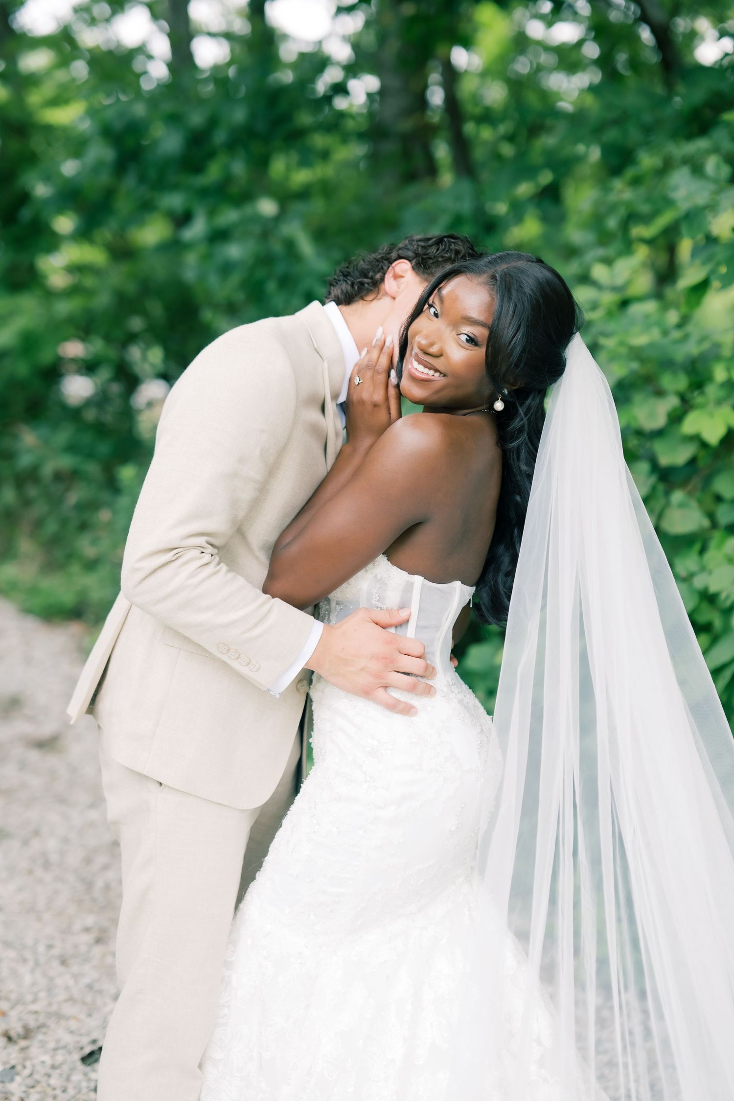 Groom in beige suit embraces bride in white lace mermaid gown and veil among lush green trees.