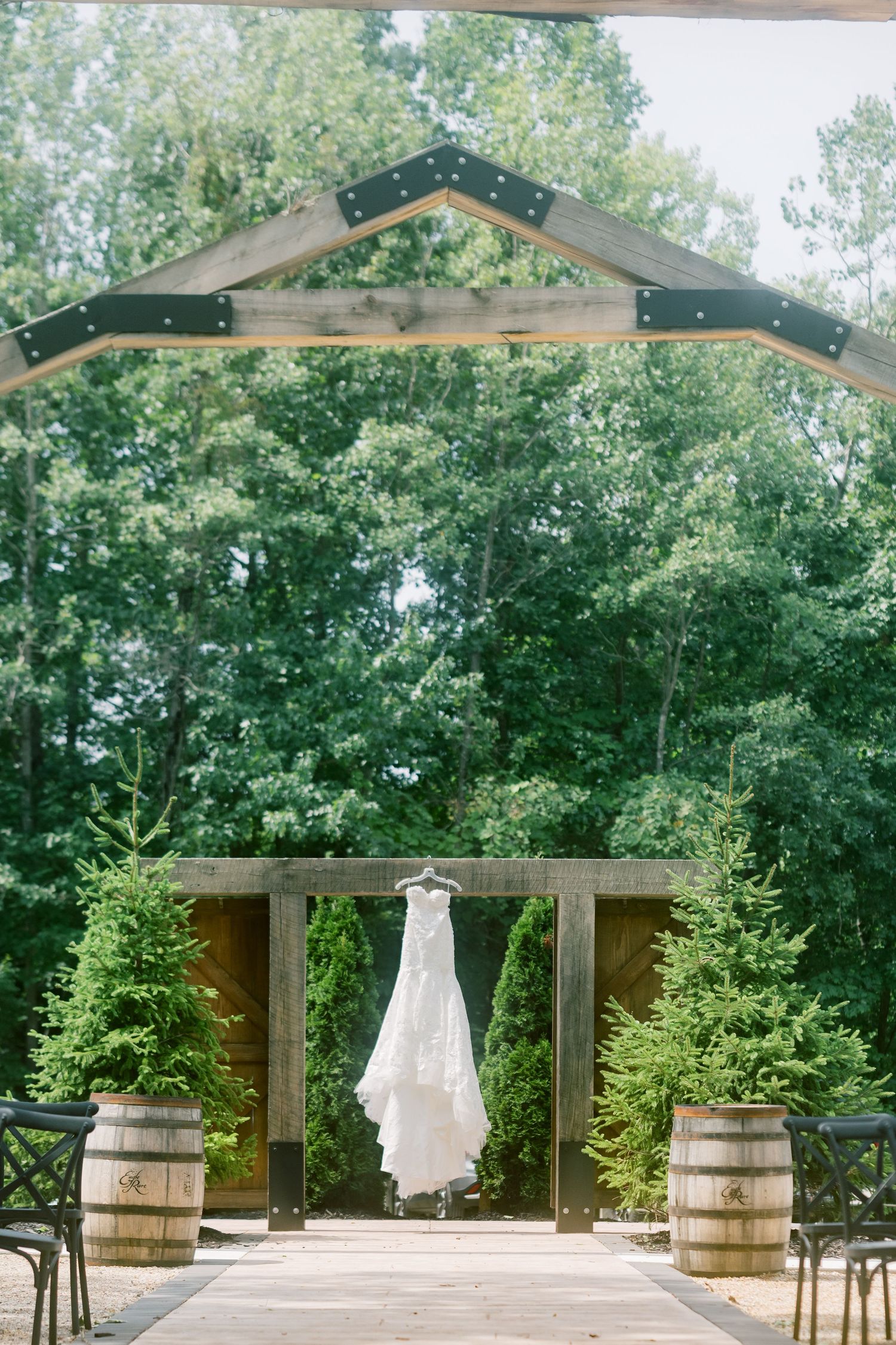 White wedding dress hanging from a rustic wooden arch surrounded by evergreen trees and barrel planters at an outdoor venue.