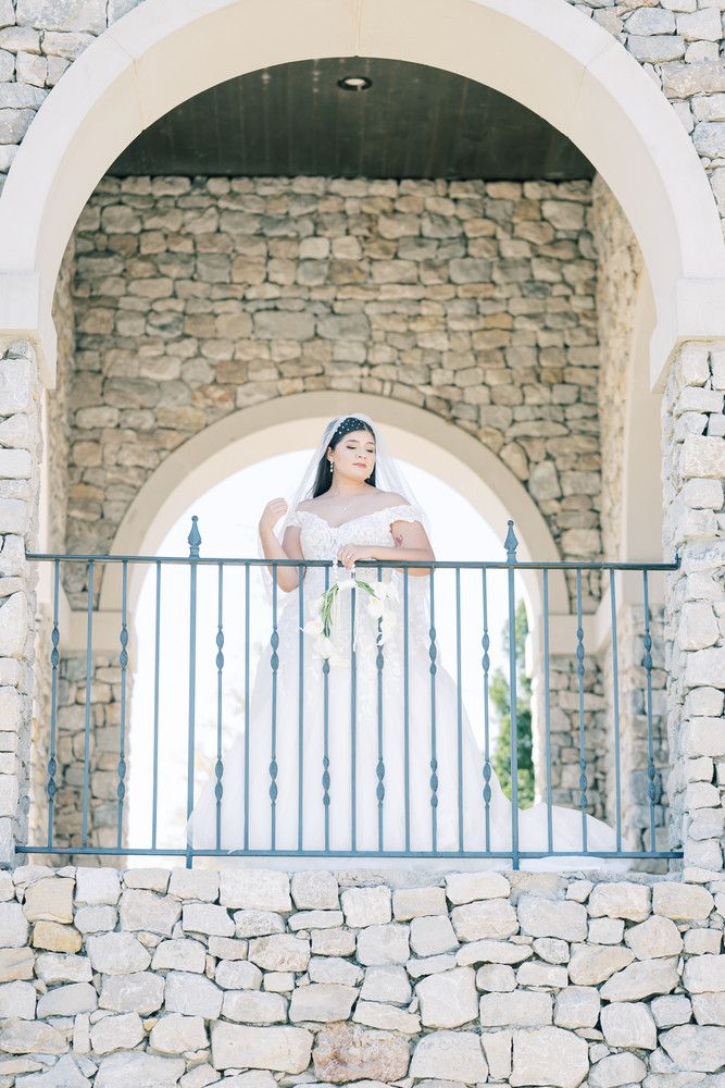 Bride in white dress stands in stone archway with decorative metal railing at scenic venue.