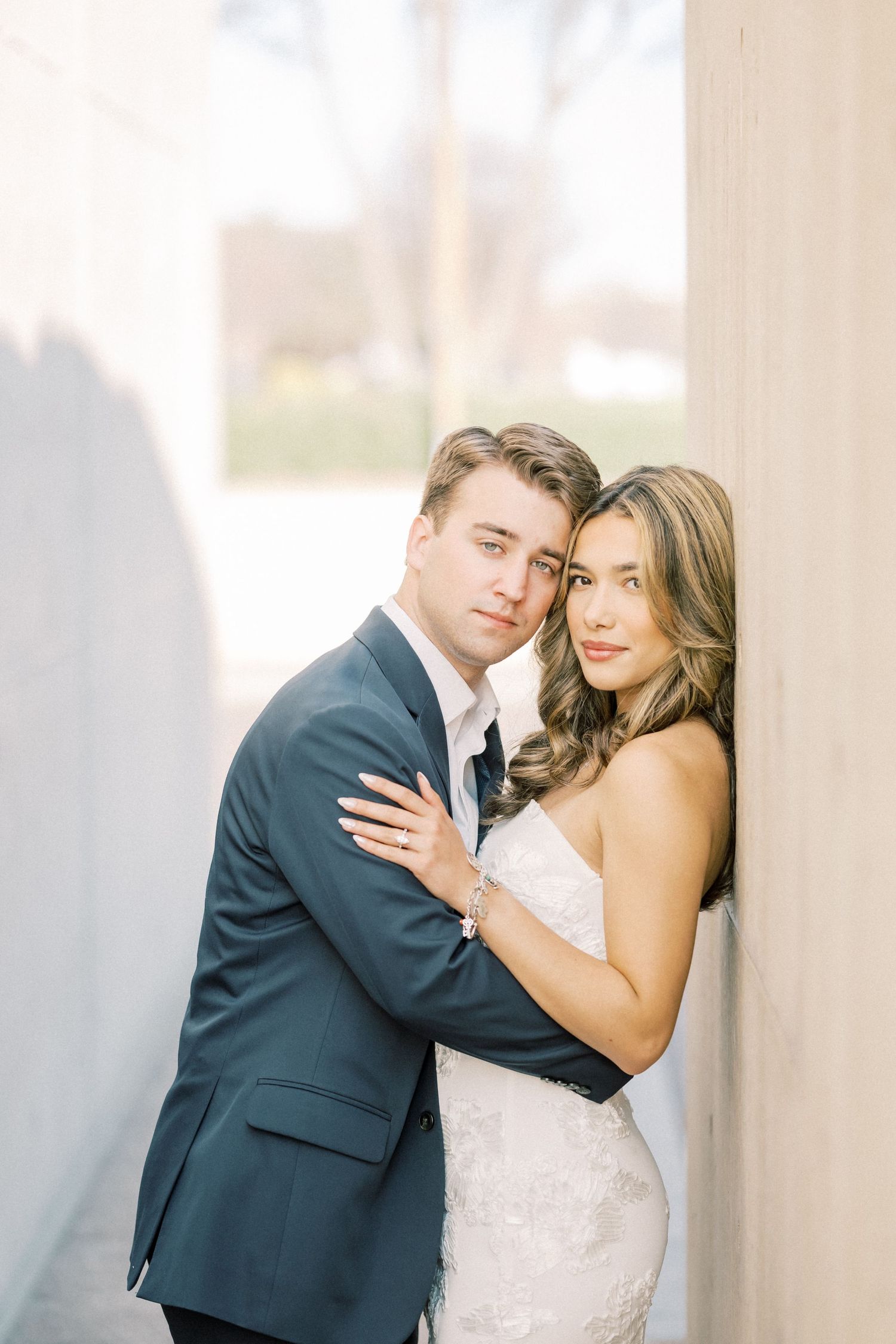 A romantic wedding couple embraces in front of white columns in elegant formal attire.