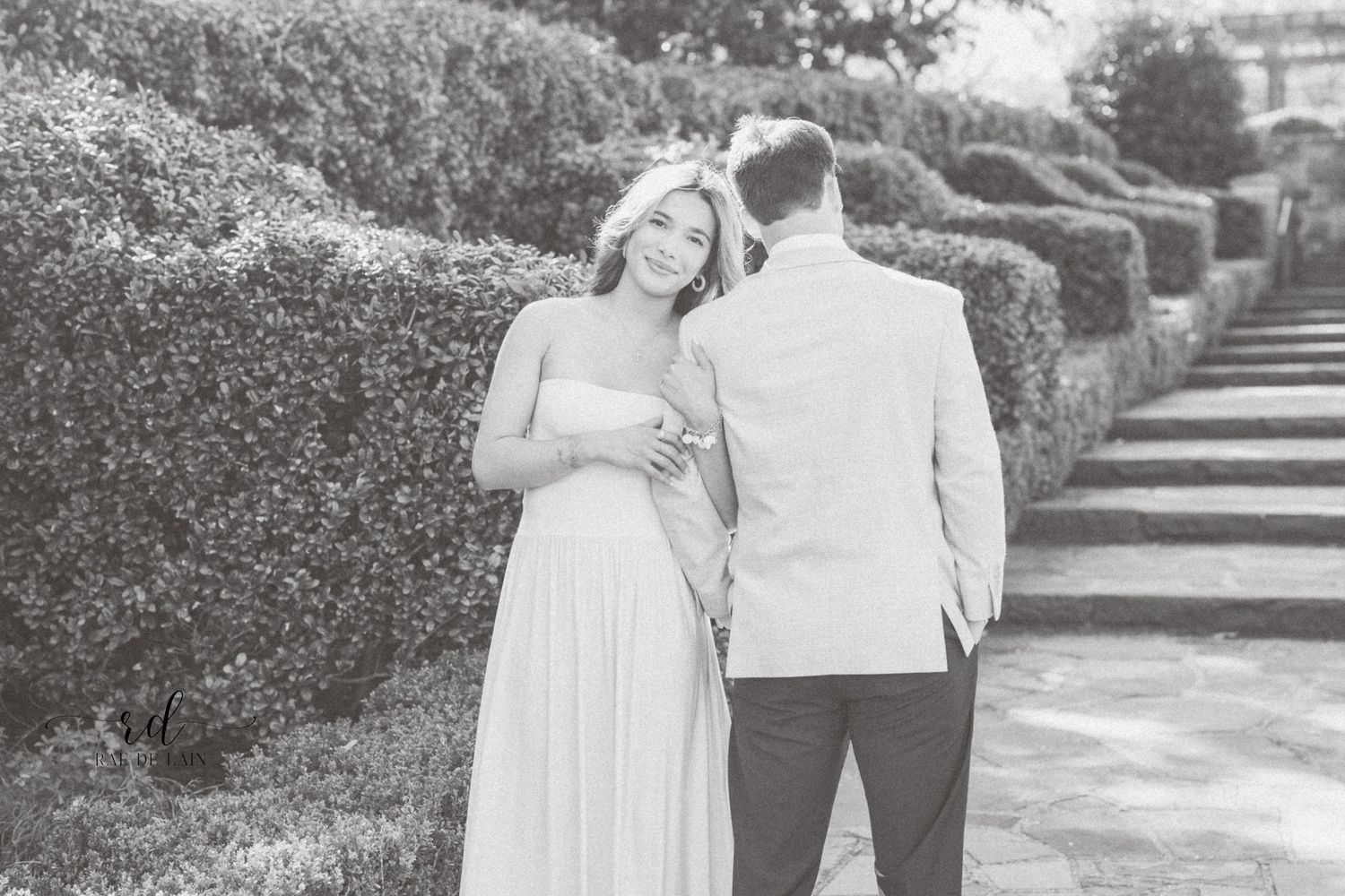 A romantic black and white photo of a couple in formal attire standing together in a garden with manicured hedges.