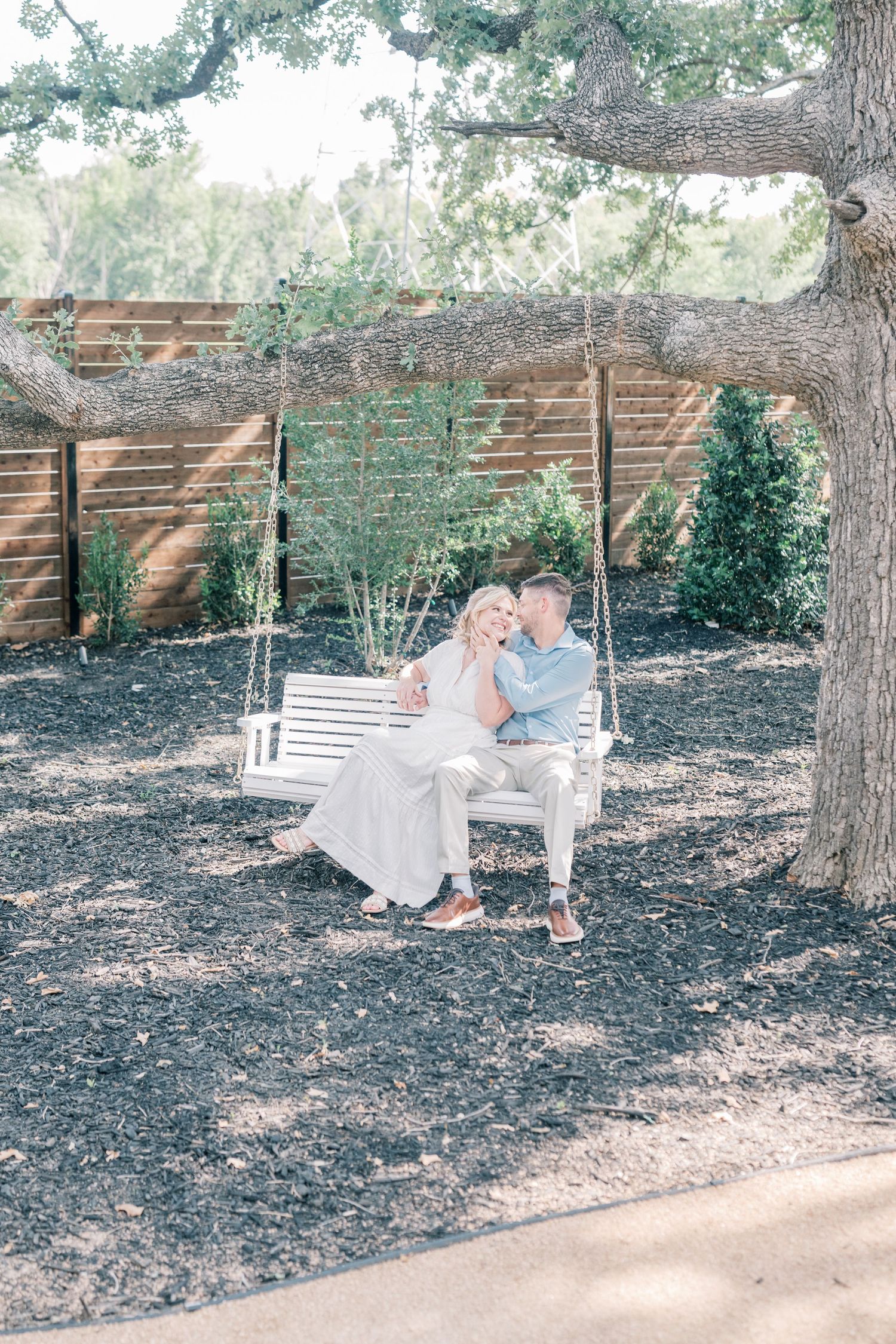 Couple sharing a tender kiss on a white wooden swing hanging from a large oak tree in a garden setting.