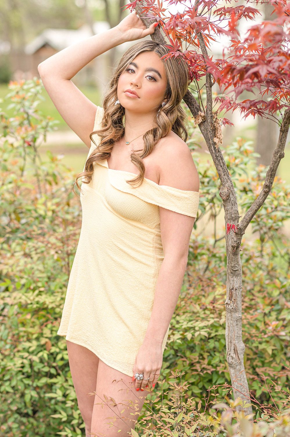 Young woman in yellow off-shoulder mini dress poses near red Japanese maple tree in lush garden setting.