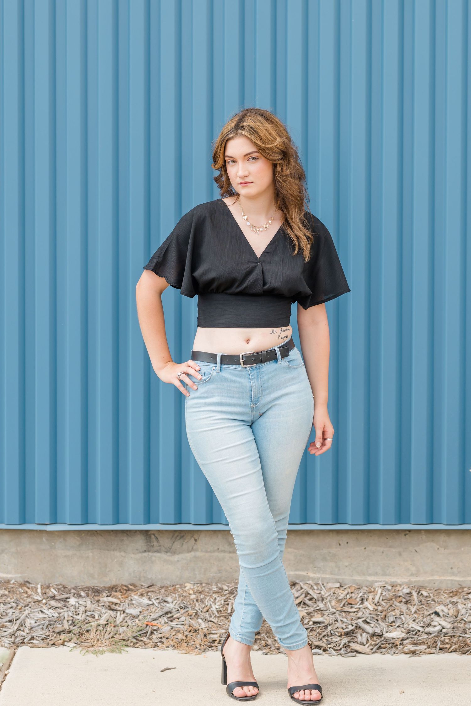 Young woman in black crop top and light blue skinny jeans posing confidently against a blue corrugated metal wall.
