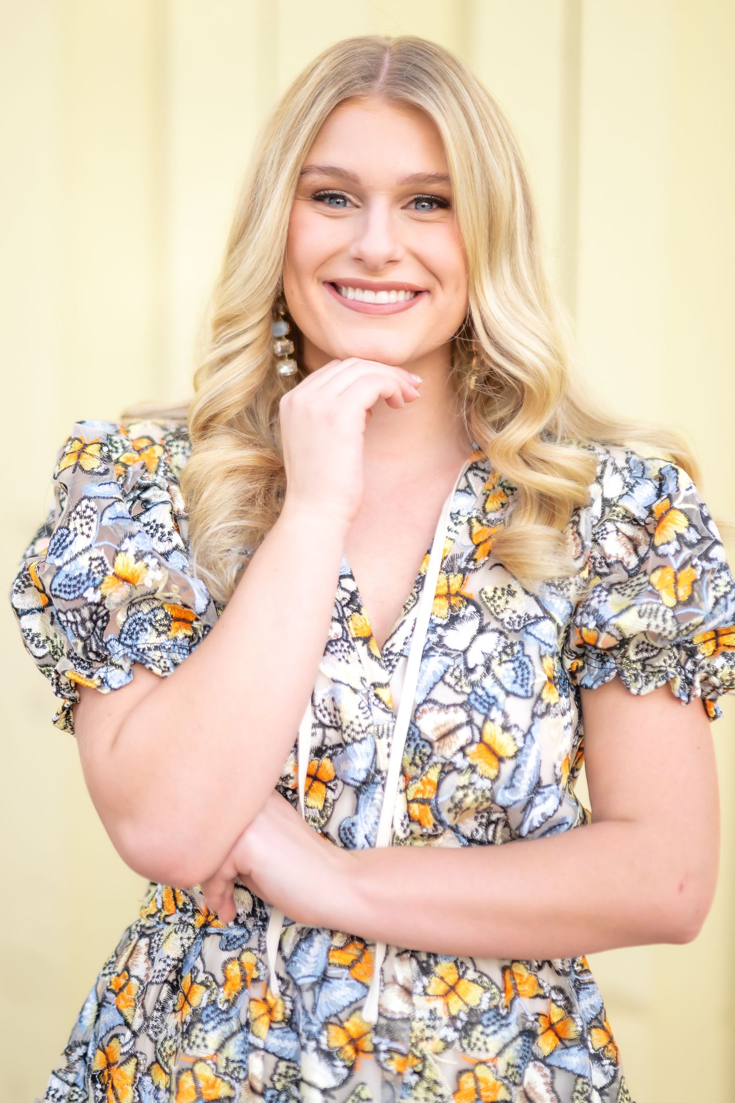 Smiling blonde woman in floral butterfly dress posing with hand on chin against yellow background.