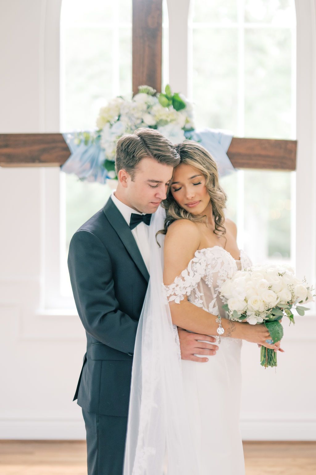 Bride and groom share tender moment in white chapel, she holds white rose bouquet in lace off-shoulder gown.