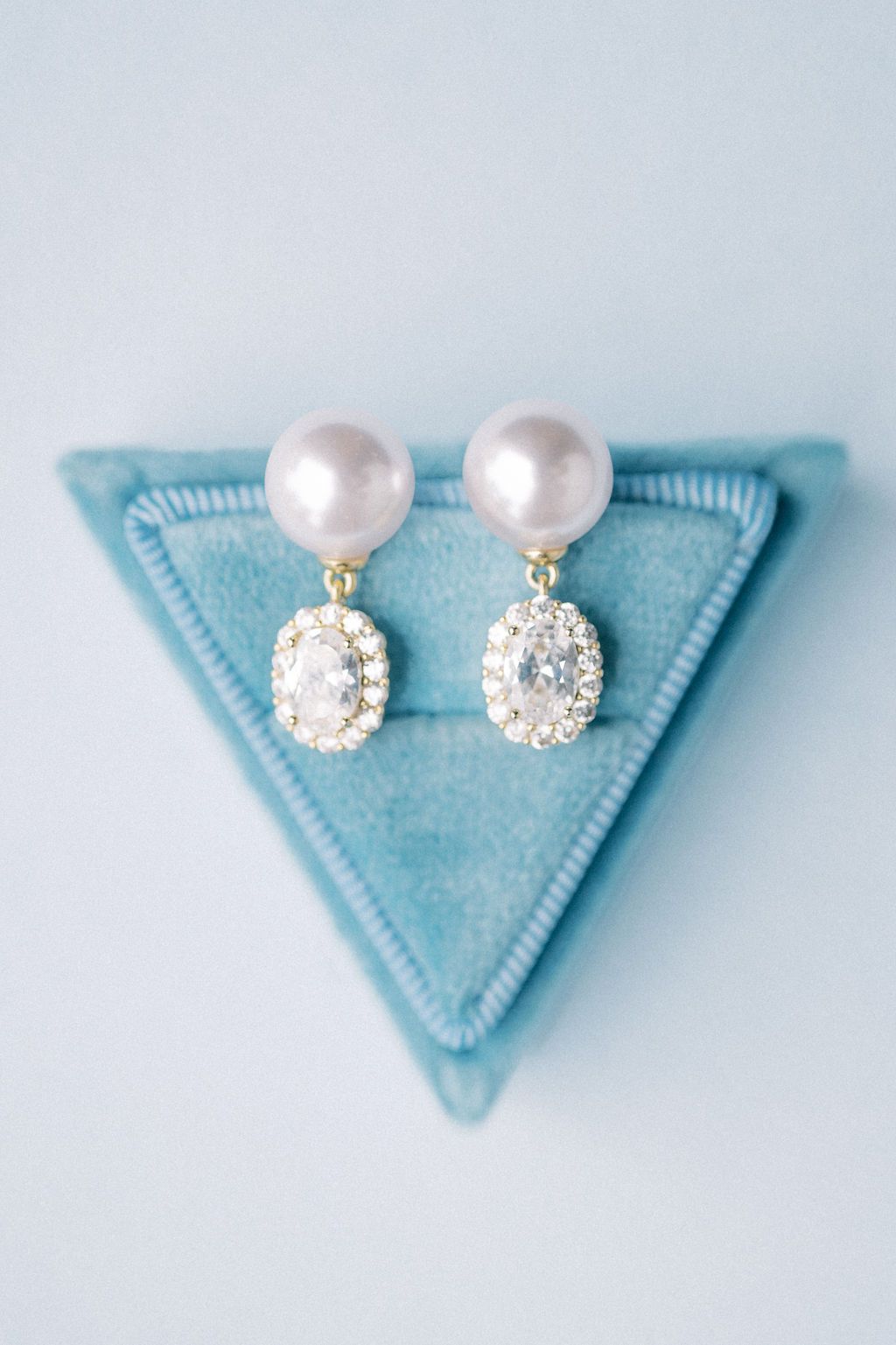 Elegant pearl and diamond halo drop earrings displayed on a triangular blue velvet jewelry box.