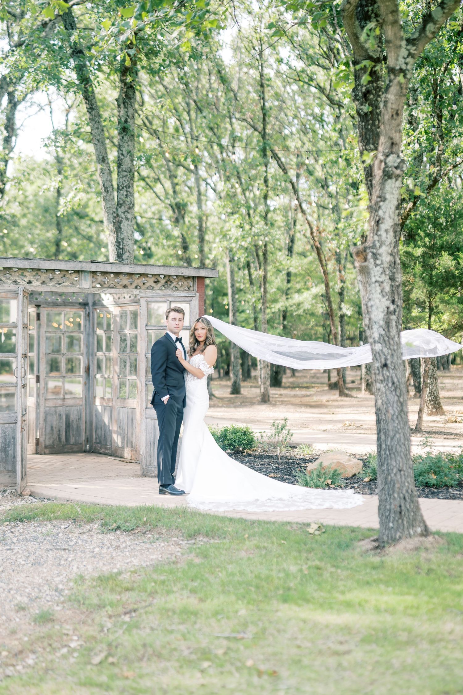 Bride and groom posing outdoors near rustic wooden doors with veil flowing in the wind among lush trees.