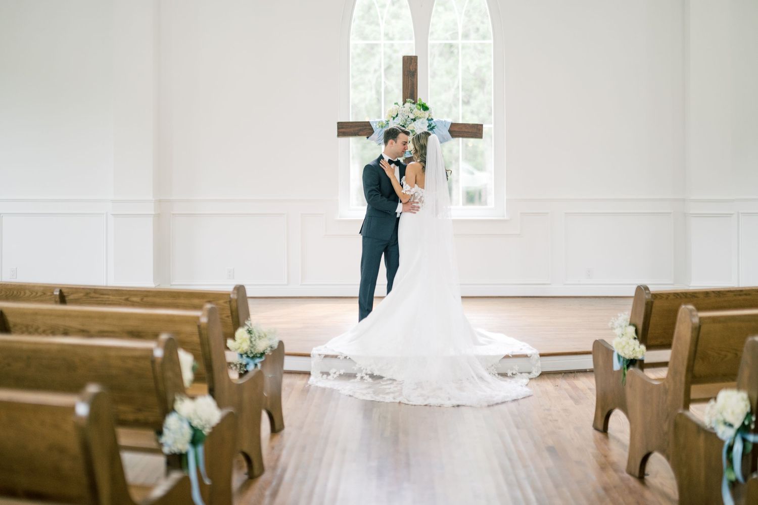 Bride and groom stand together at chapel altar with wooden cross, surrounded by white pews and flowers.
