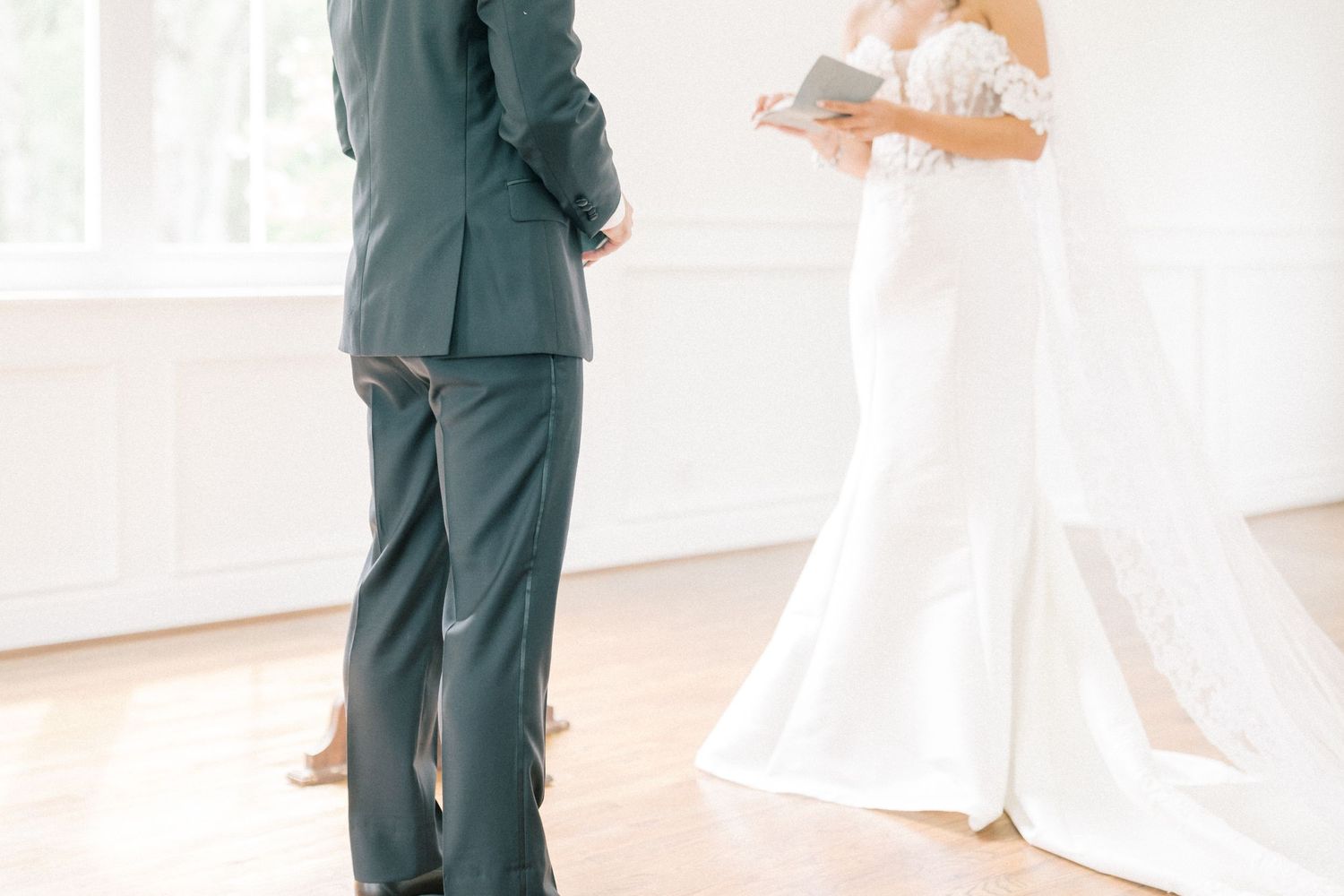 Bride in white gown reads vows to groom in gray suit during intimate indoor wedding ceremony.