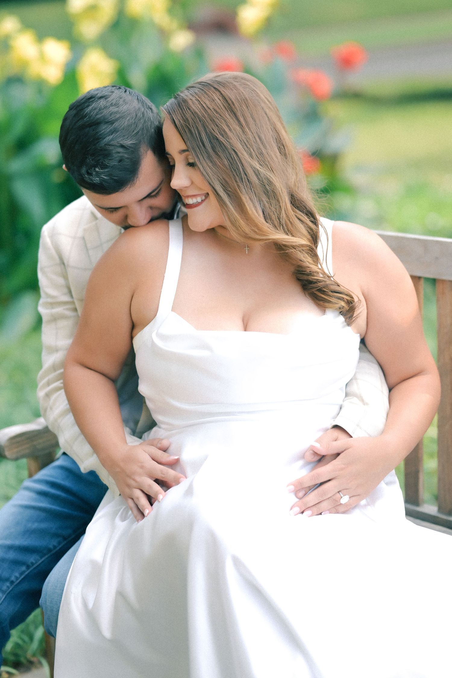 Smiling woman in a white dress sits on a garden bench as a man kisses her shoulder and rests his hands on her waist.