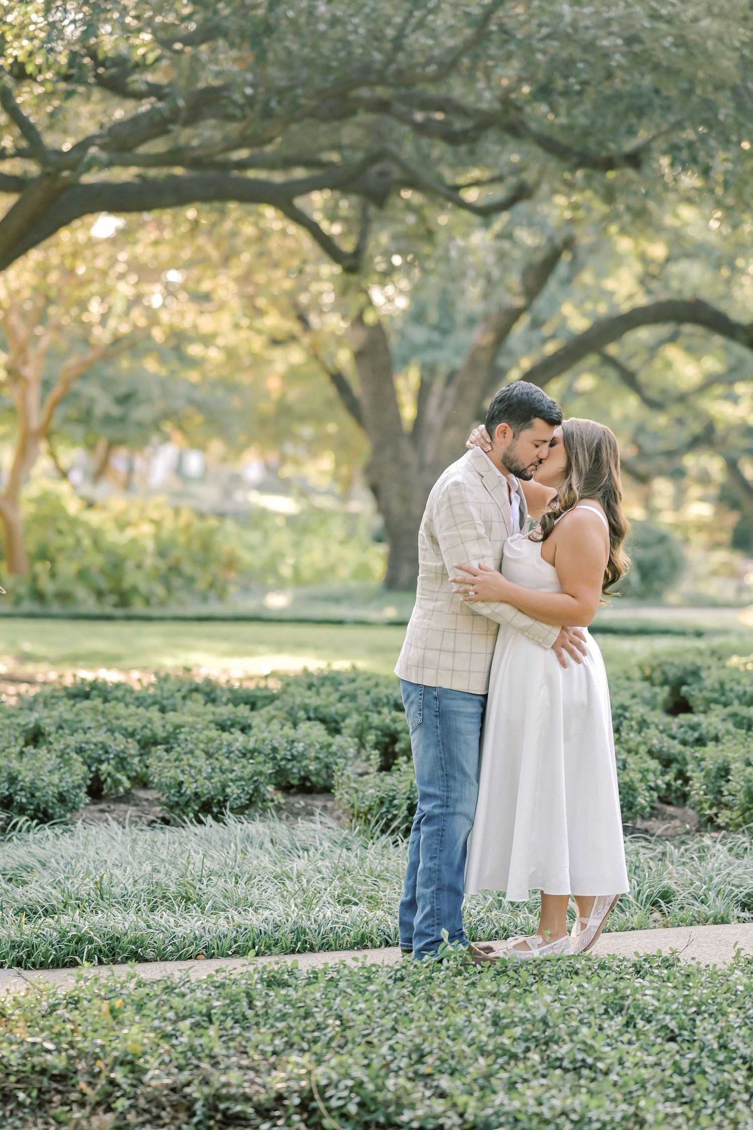Engaged couple share a kiss on a garden path beneath large oak trees, she in a white dress and he in a plaid blazer.