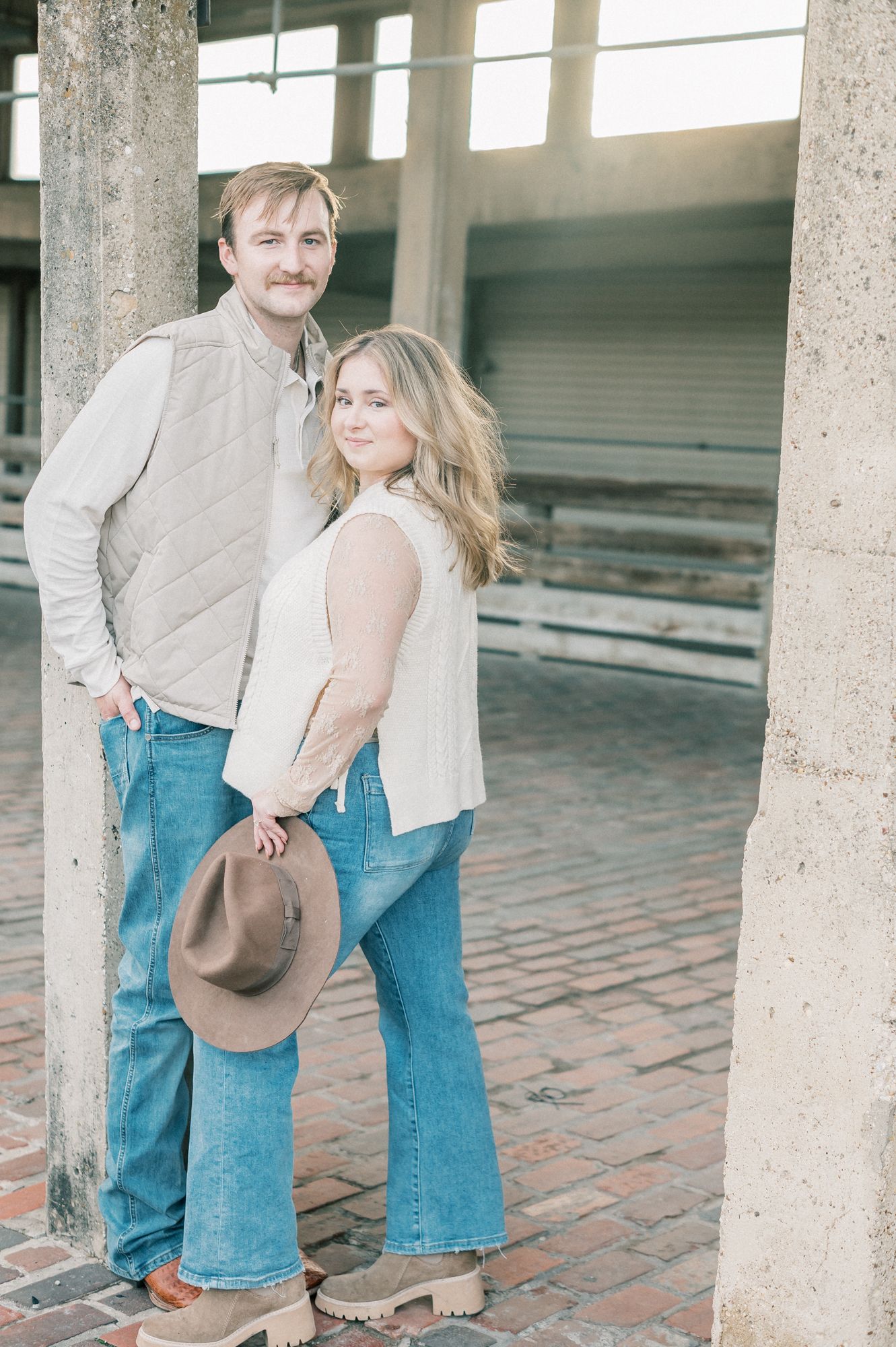 A couple in denim and light clothing poses playfully between concrete pillars on brick pavement.