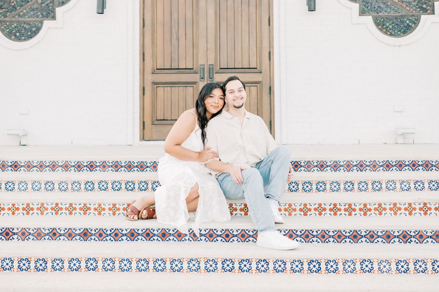 Couple in light blue and white outfits pose together against a decorative tile wall background.