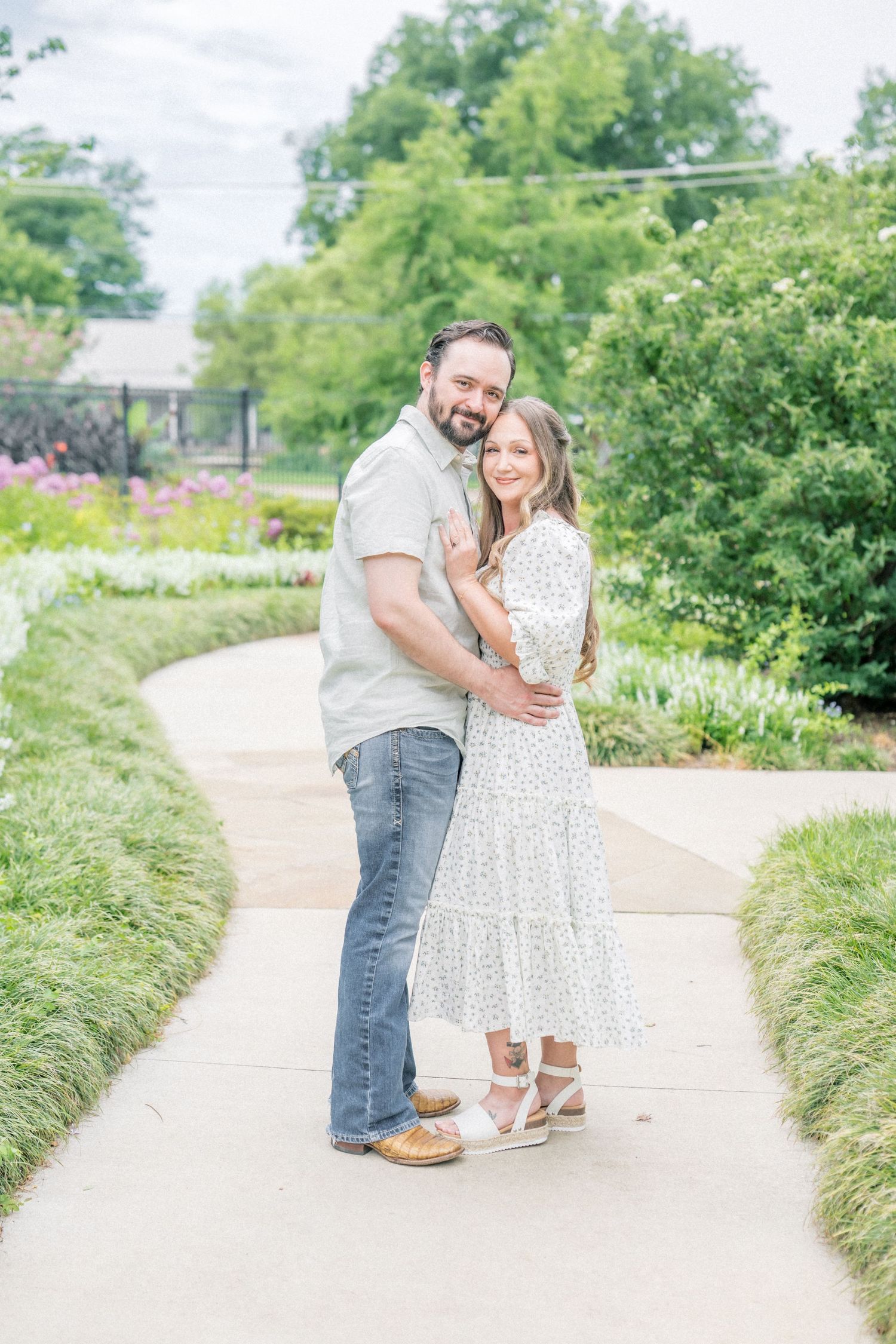 A couple embraces on a garden path, the man in a gray shirt and jeans, the woman in a white floral midi dress.