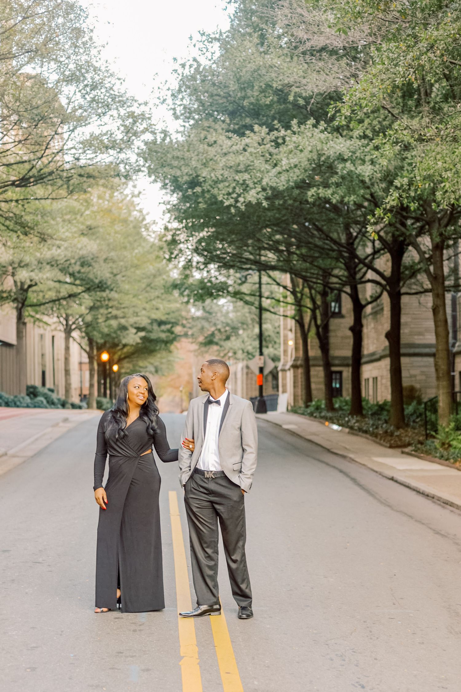 Elegant couple in formal attire standing on a tree-lined street, woman in black jumpsuit and man in gray suit.