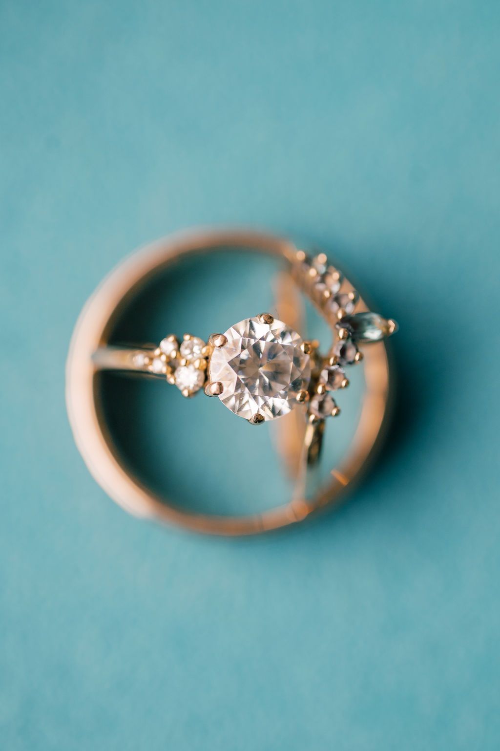 Gold engagement ring with round diamond center stone and cluster accents displayed on teal background.