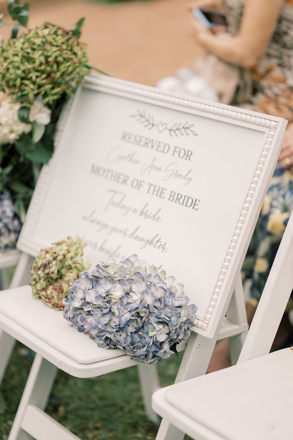 White framed reserved sign for Mother of the Bride adorned with blue hydrangeas on white chairs at outdoor wedding.