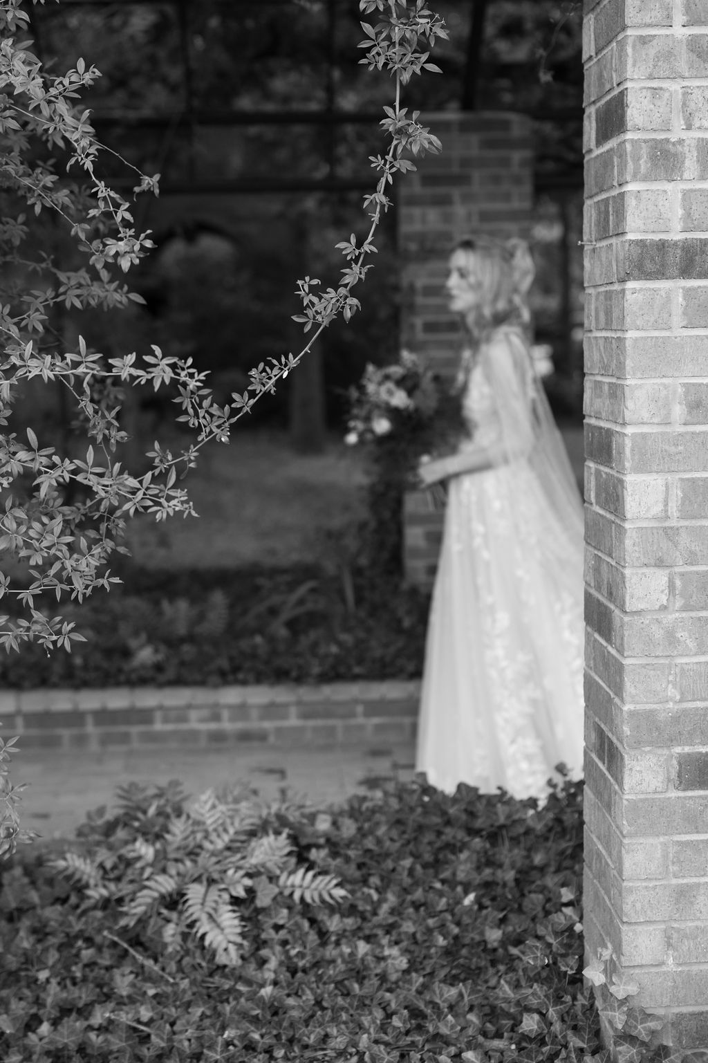 Black and white photo of a bride in a lace gown holding a bouquet, standing near a brick pillar surrounded by lush greenery.