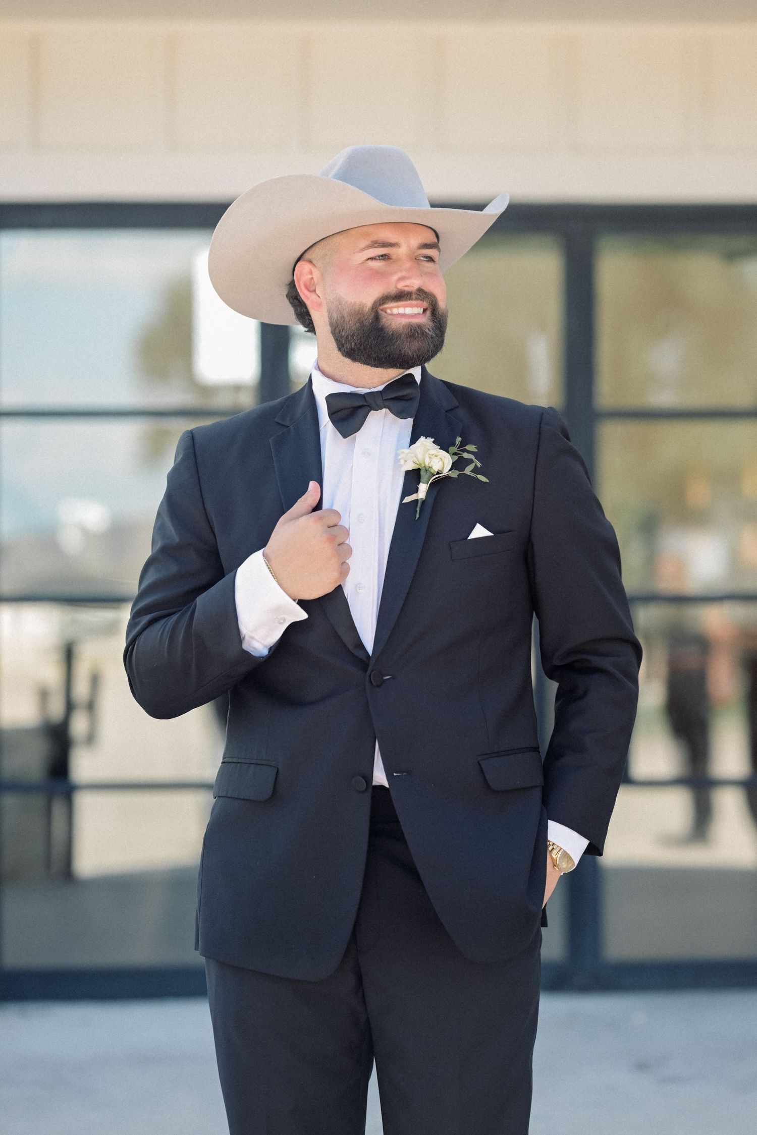 Smiling groom in black tuxedo with white cowboy hat and boutonniere poses outside wedding venue.