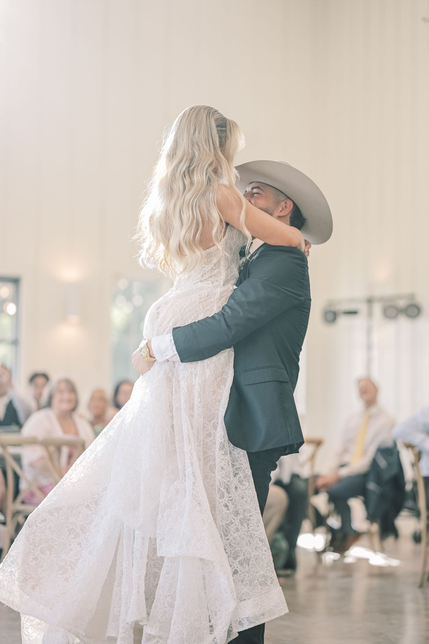 Bride and groom share first dance embrace in bright reception hall while guests watch in background.