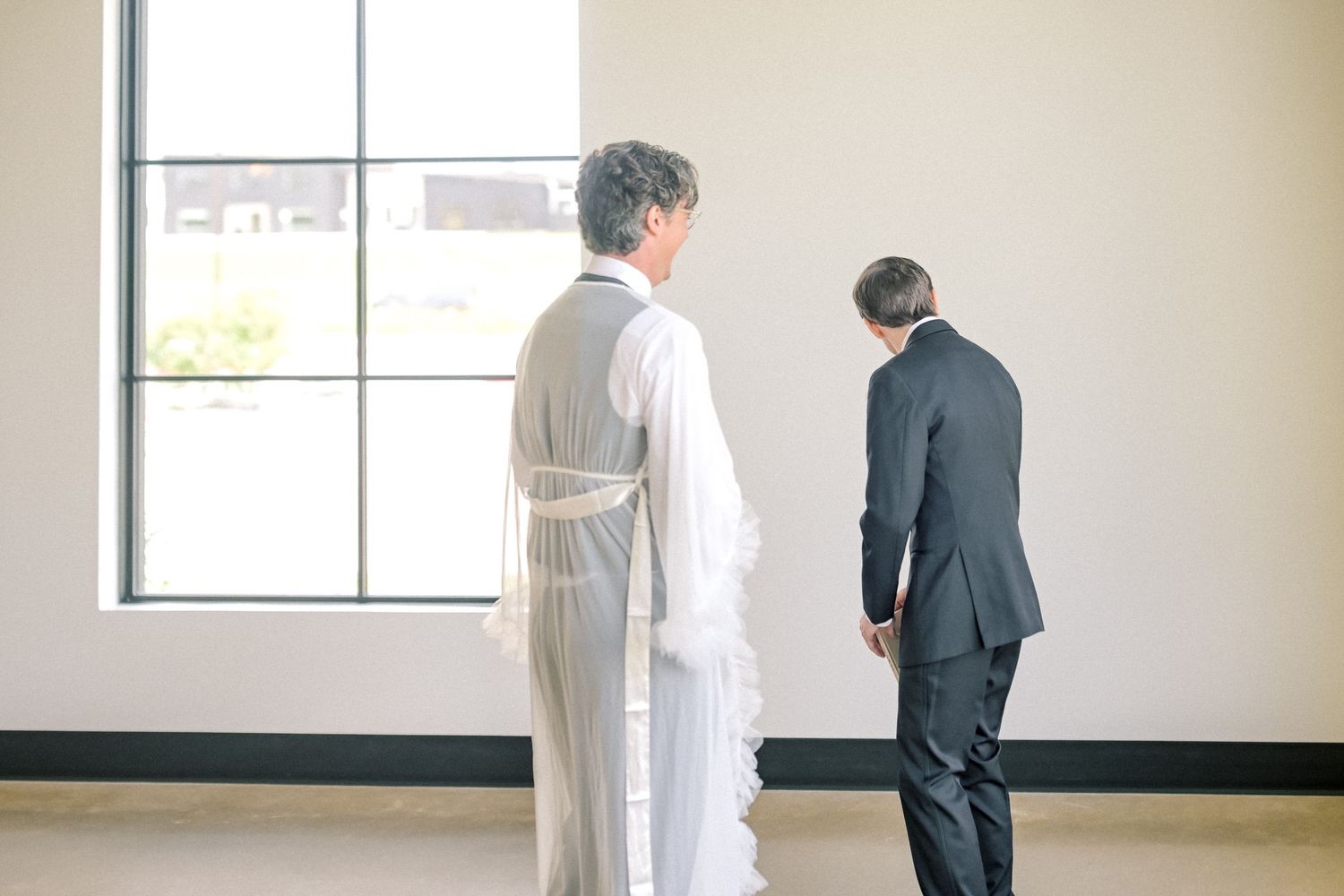 Bride and groom share a quiet first look moment standing by a large window in a bright venue.