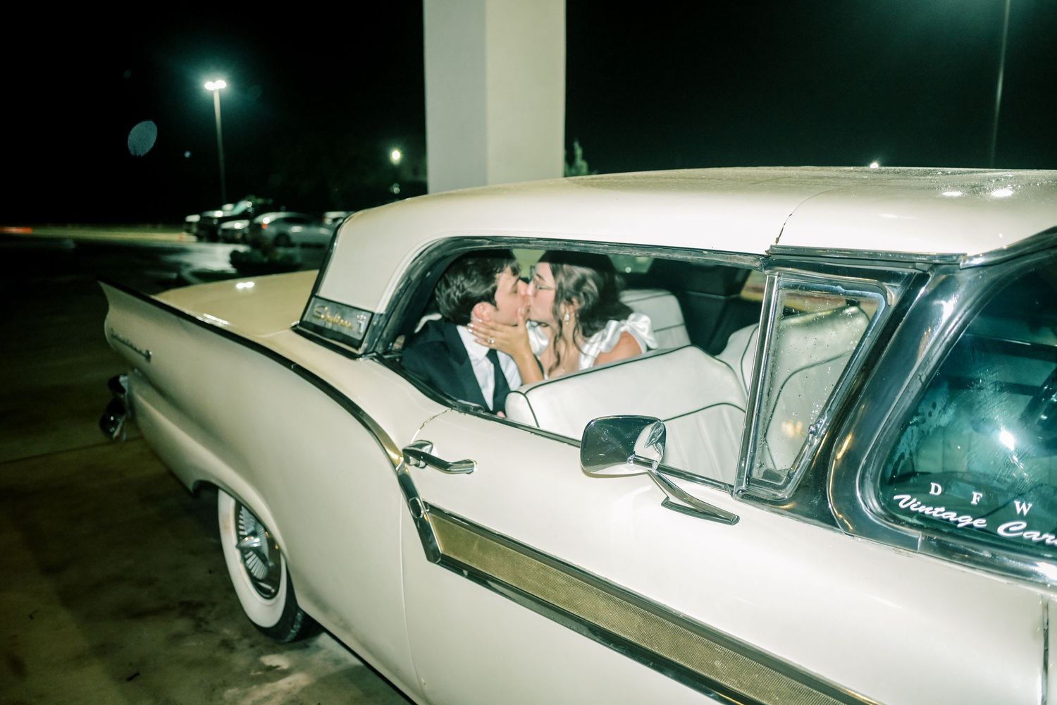 Bride and groom sharing a kiss inside a vintage cream colored car at night.