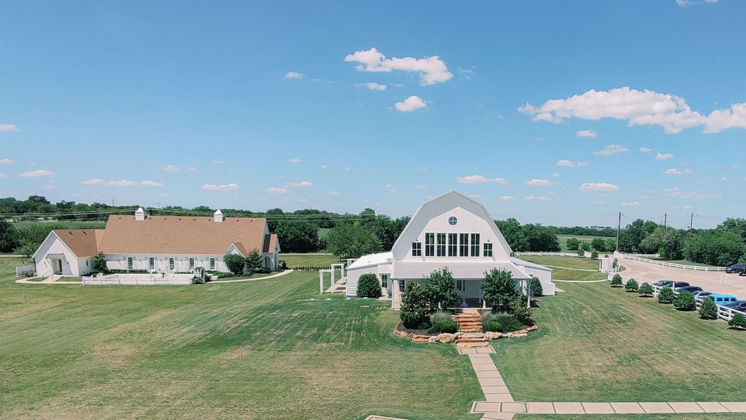 Aerial view of a white barn wedding venue with large windows, surrounded by lush green lawns under a bright blue sky.