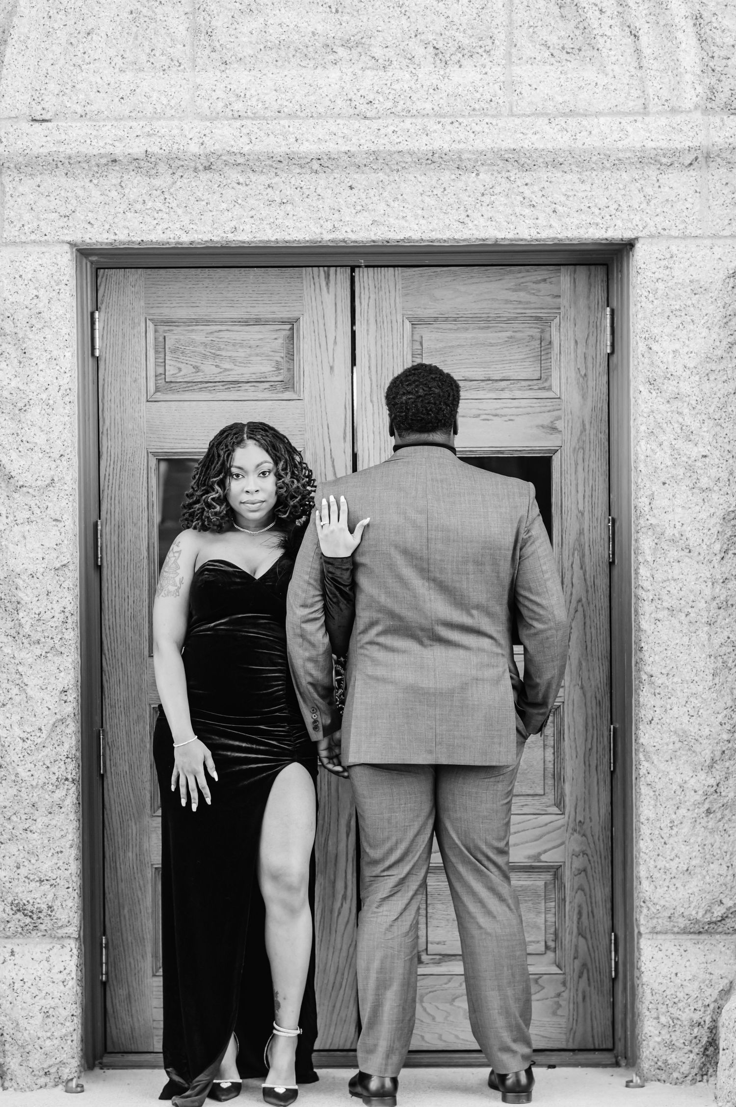 Black and white photo of a couple in formal wear posing by wooden doors, woman in black velvet gown, man in gray suit.