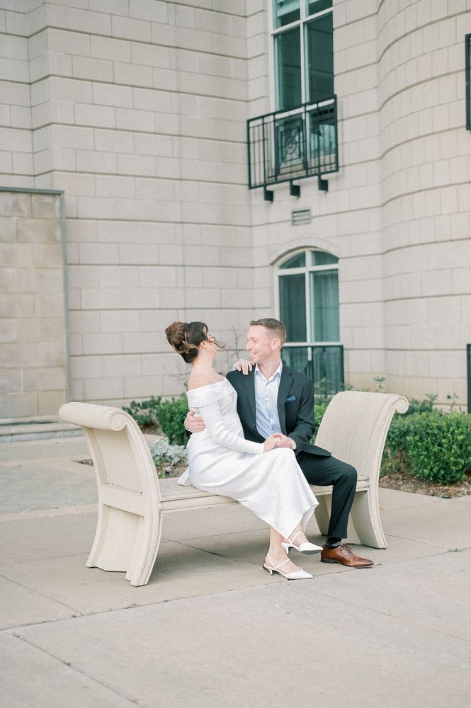 Couple in wedding attire sitting on a bench outside a stone building, sharing a romantic moment.