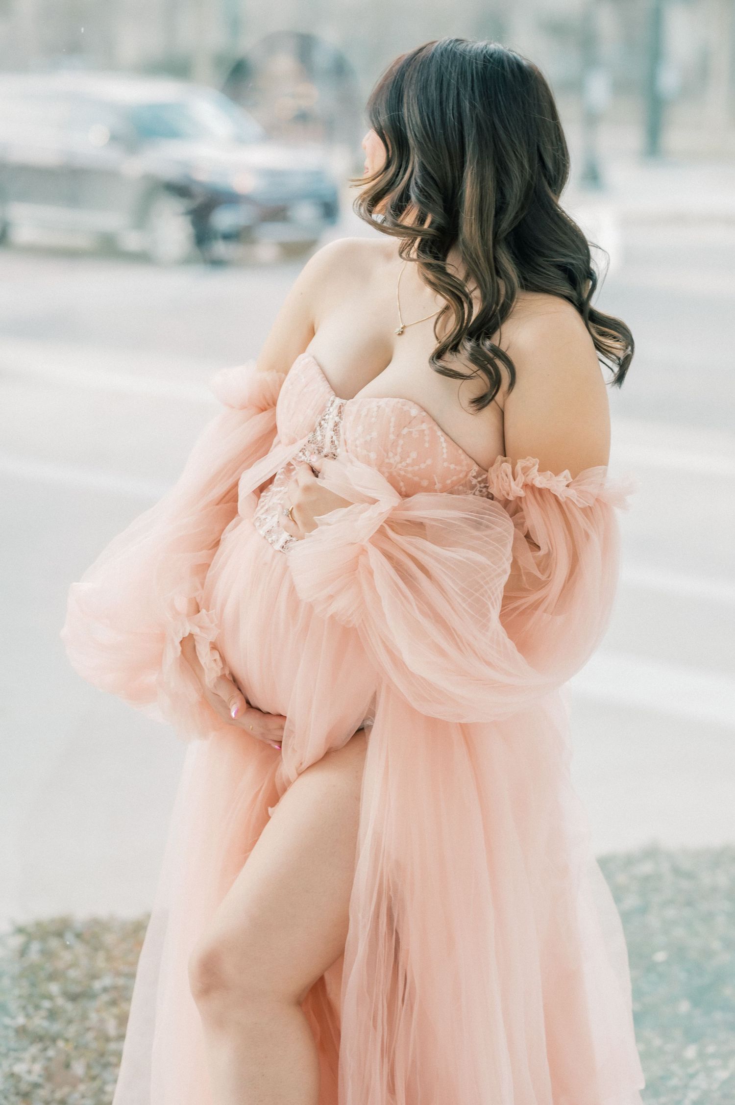 Pregnant woman in pink tulle maternity gown poses outdoors showing baby bump.