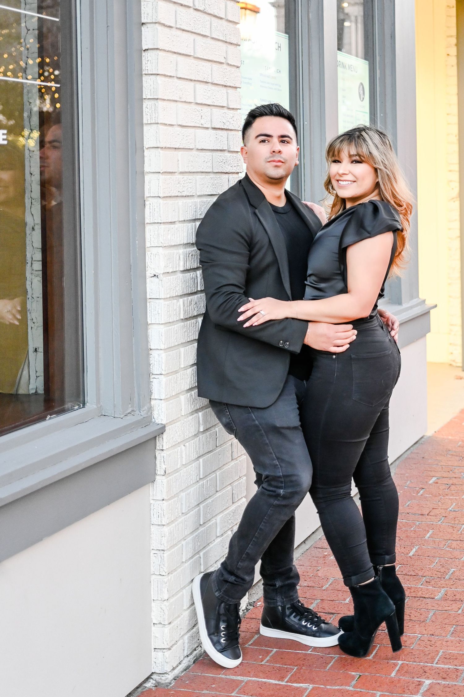 Stylish couple in all-black outfits posing against a white brick wall in an urban setting.