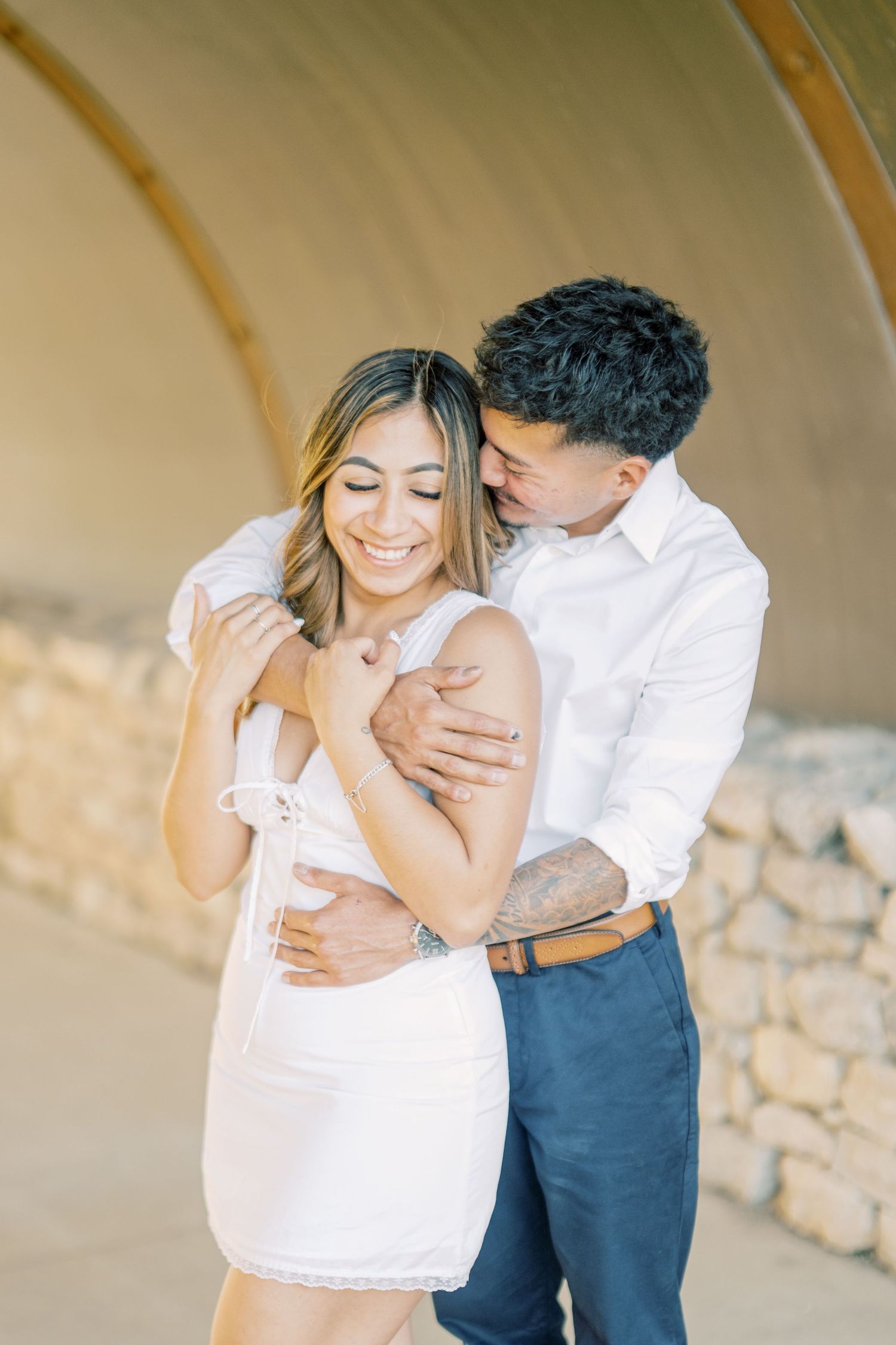 Happy couple sharing a tender moment under a stone arch, woman laughing in white mini dress.