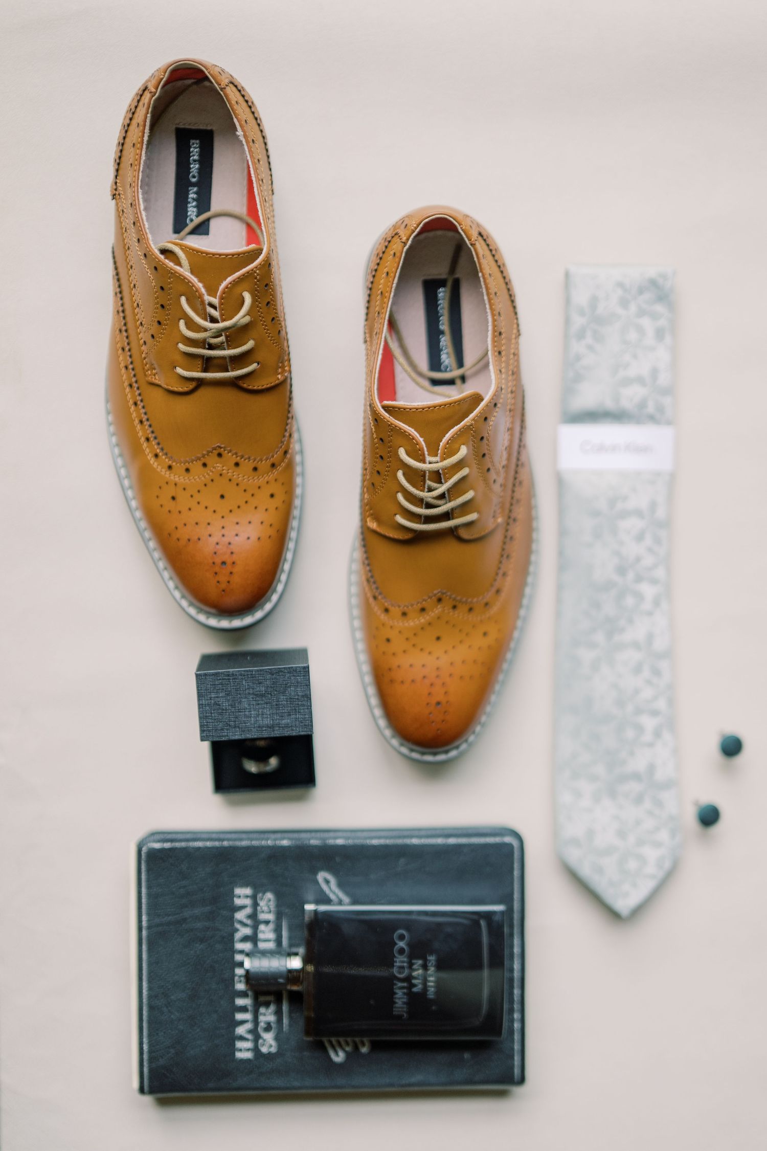 Groom's wedding accessories flat lay featuring tan brogue shoes, grey floral tie, cufflinks, cologne, and a book.