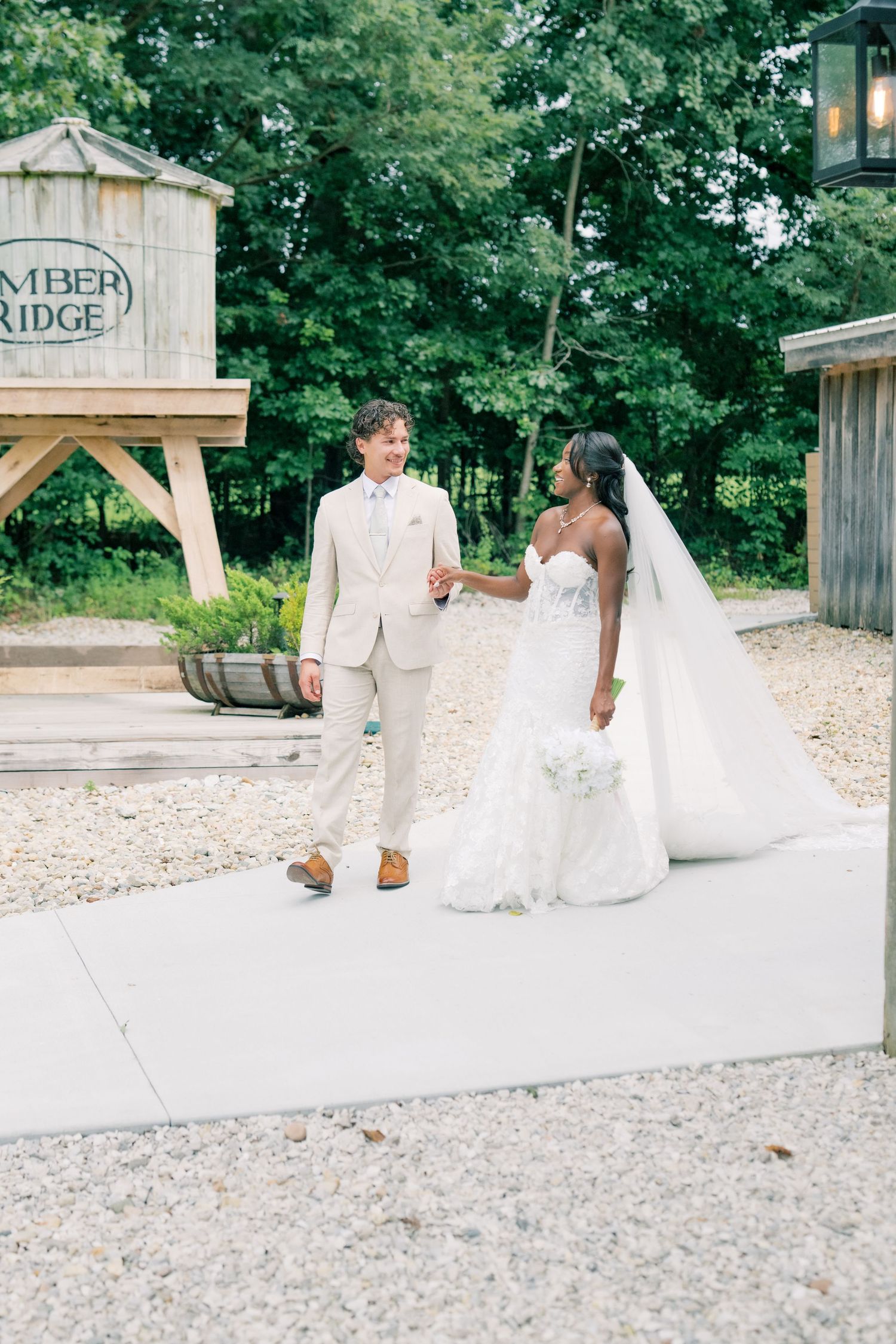Interracial couple walking hand in hand at outdoor wedding venue, groom in ivory suit, bride in lace gown with veil.