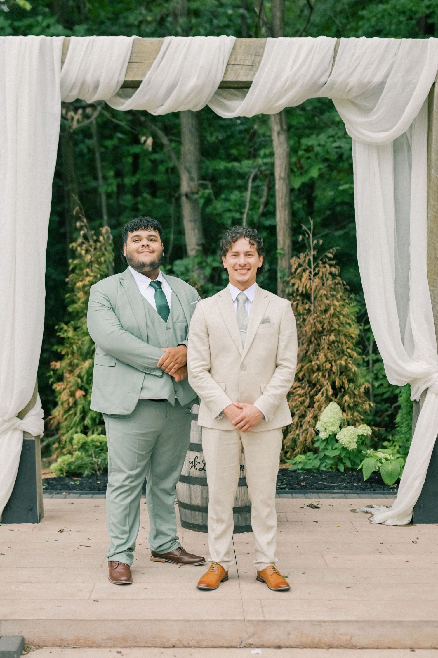 Two grooms smile under a white draped wedding arch outdoors, one in sage green suit, one in beige suit.