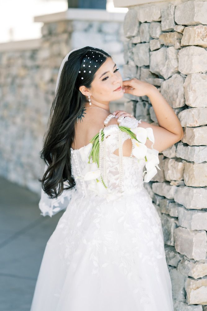Bride in white lace gown poses against stone wall with delicate pearl hair accessories and dramatic lighting.