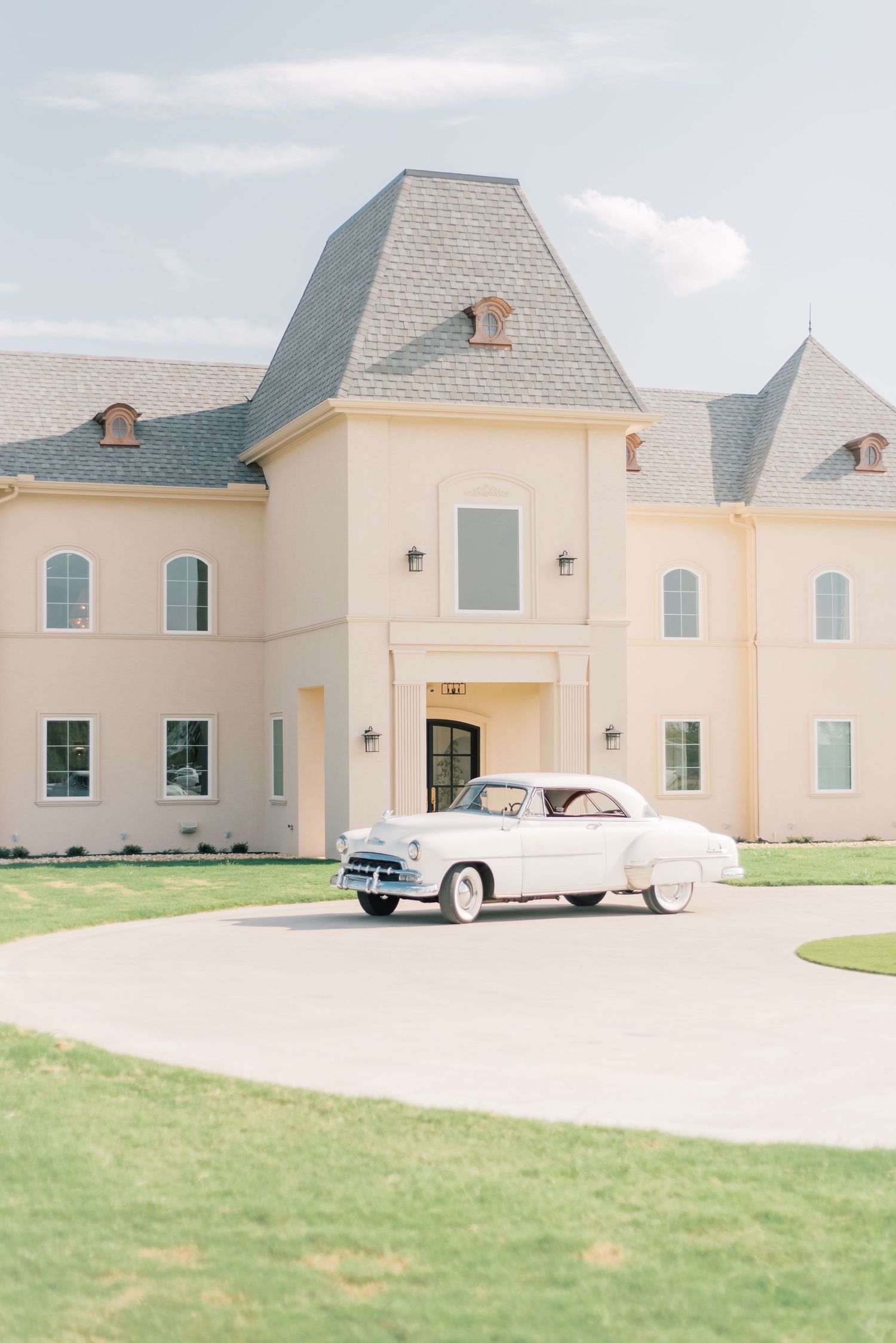 Classic white vintage car parked in front of an elegant cream-colored French chateau style mansion.