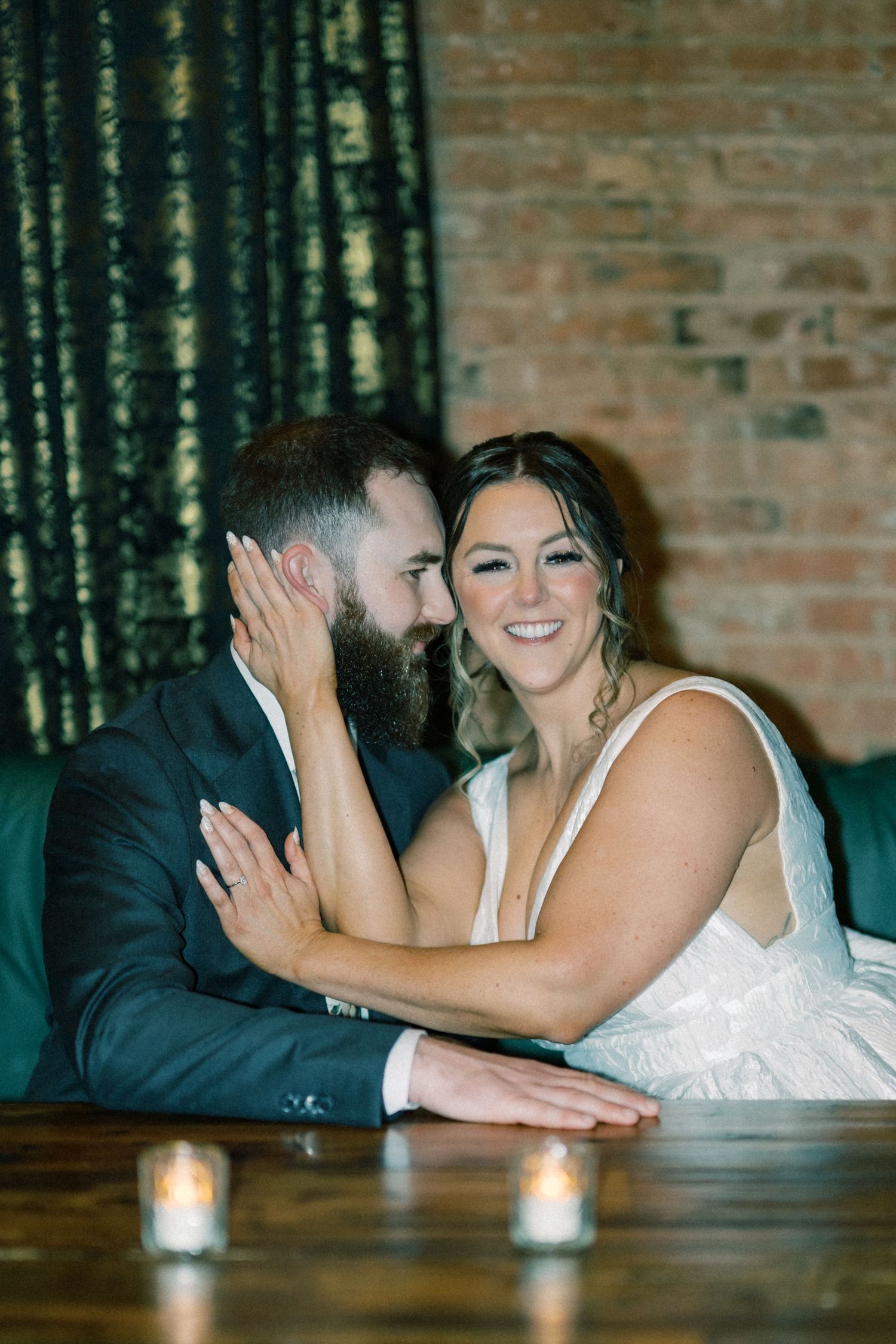 Romantic couple poses on wooden table with candlelight at rustic brick venue.