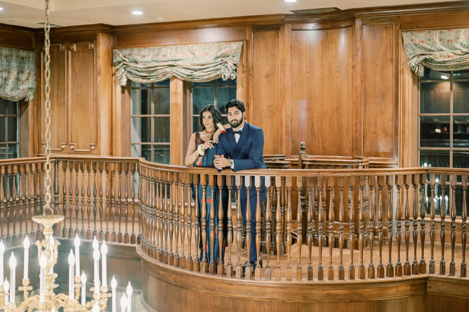 Couple stands together on elegant wooden balcony with chandelier below.