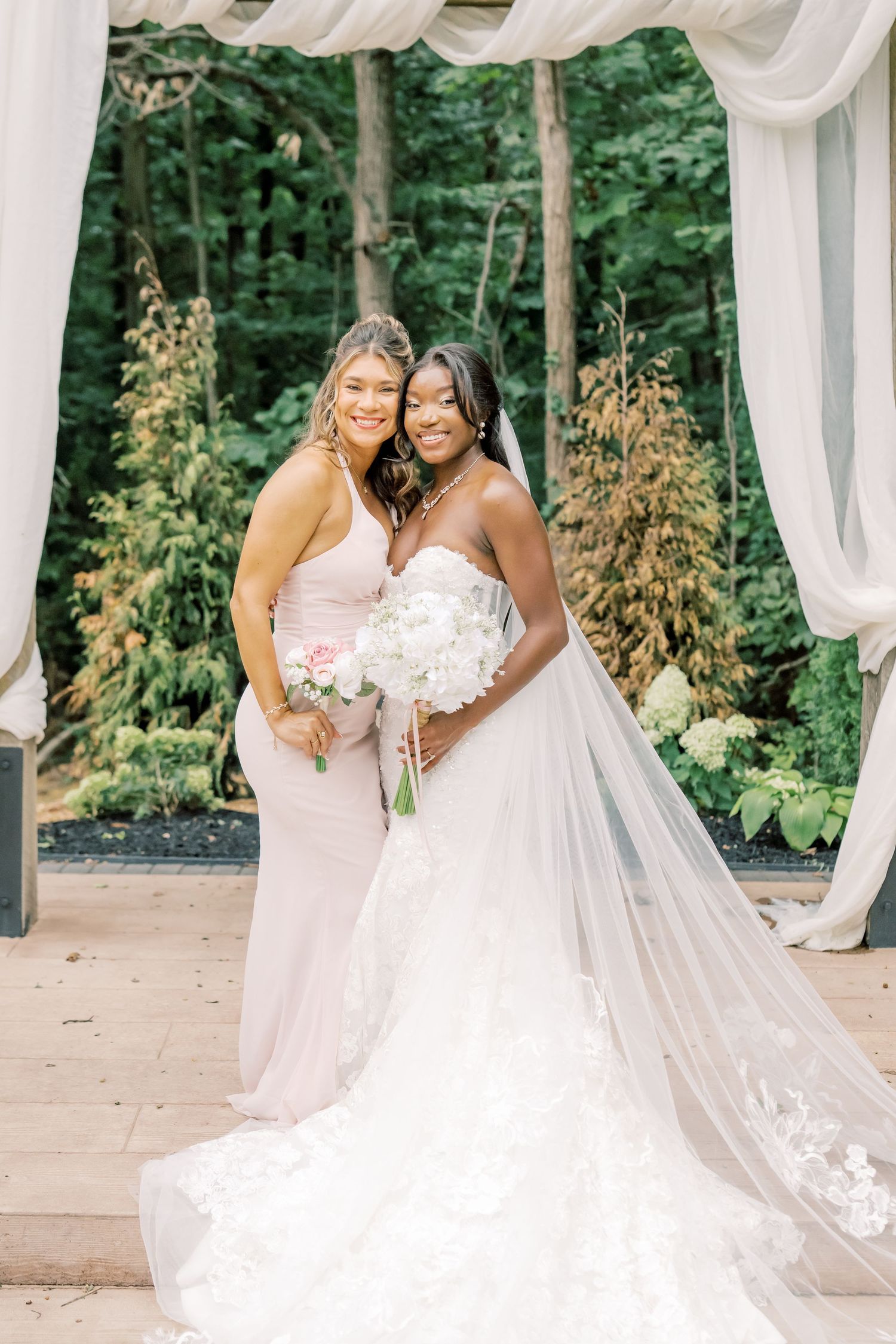 Bride in lace gown with veil and bridesmaid in blush dress pose under draped arch at outdoor wedding.