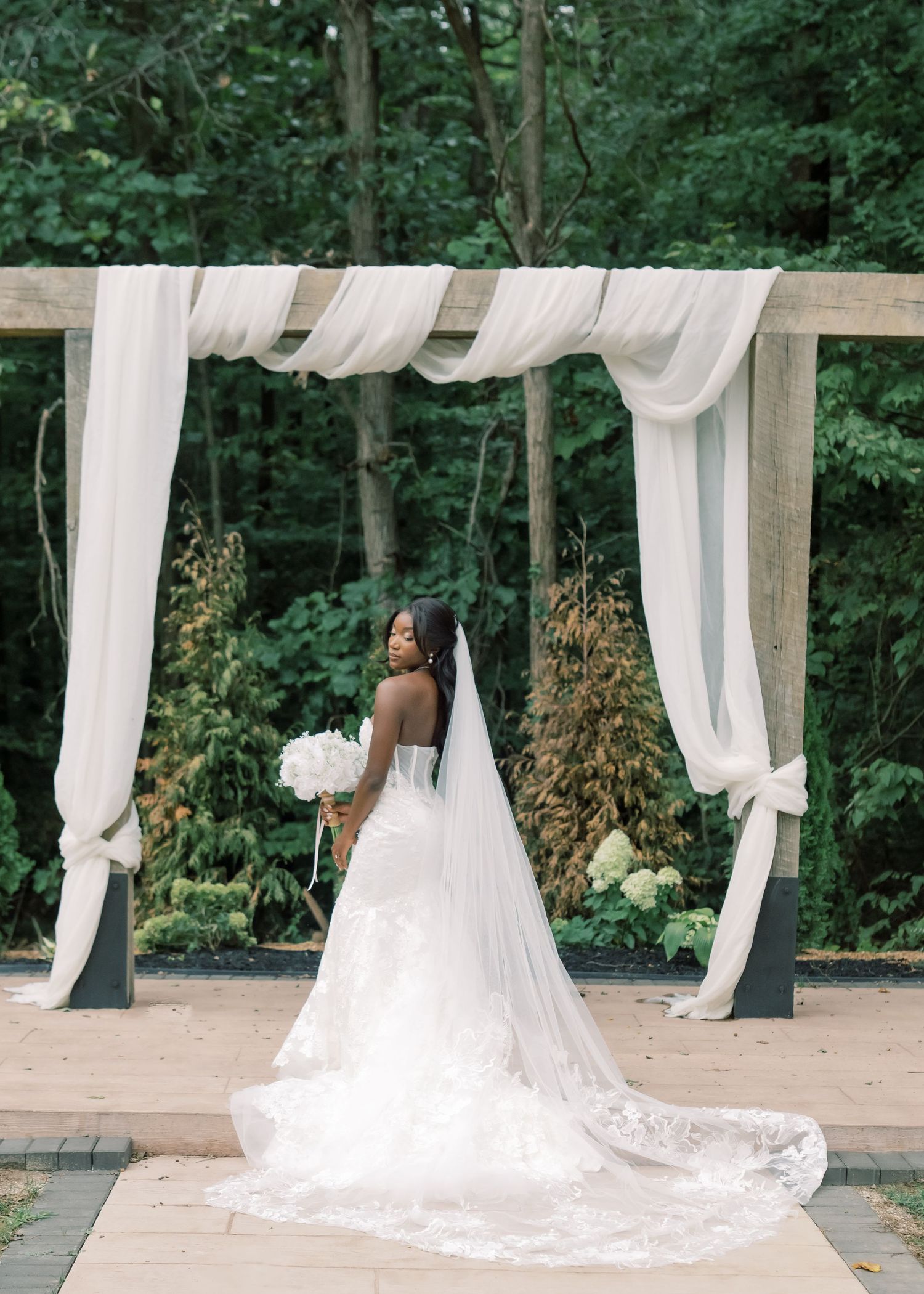 Black bride posing with back to camera under white draped wedding arch, wearing lace gown with long cathedral veil.
