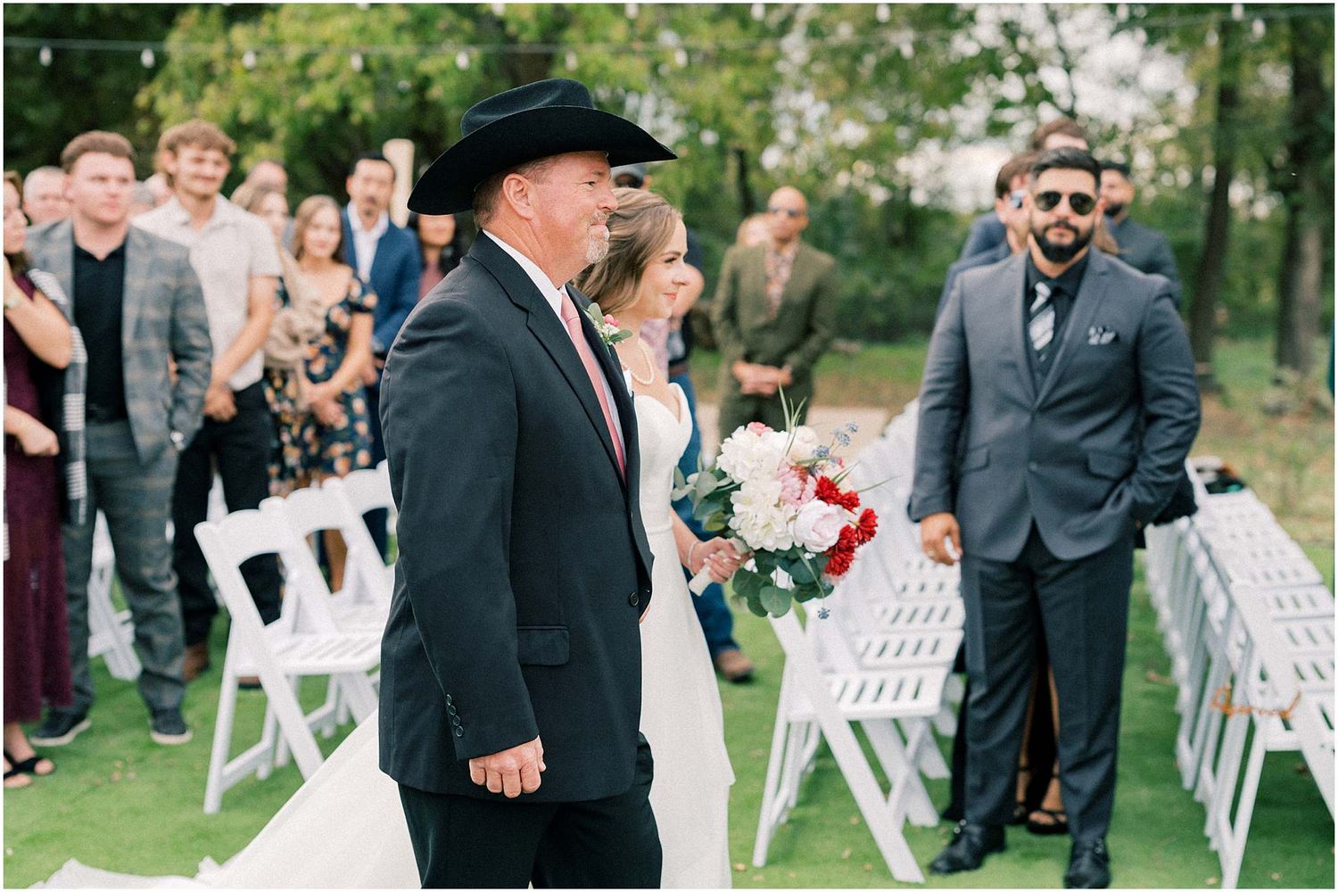 Outdoor wedding ceremony with guests seated on white chairs under trees.