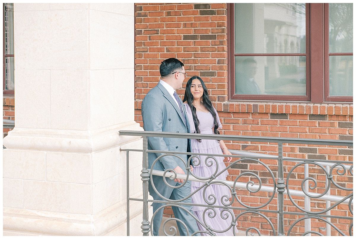 A couple poses along a brick building with decorative railings.