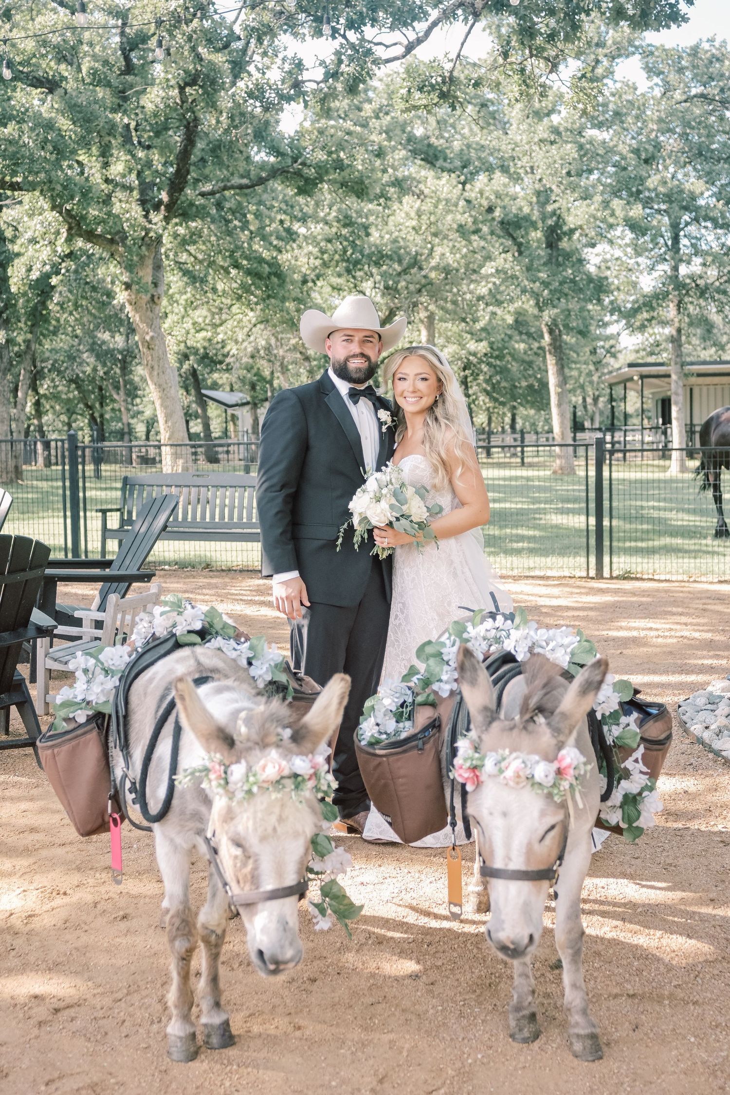 Bride and groom in cowboy hat pose with flower-adorned donkeys at rustic outdoor Texas wedding venue.